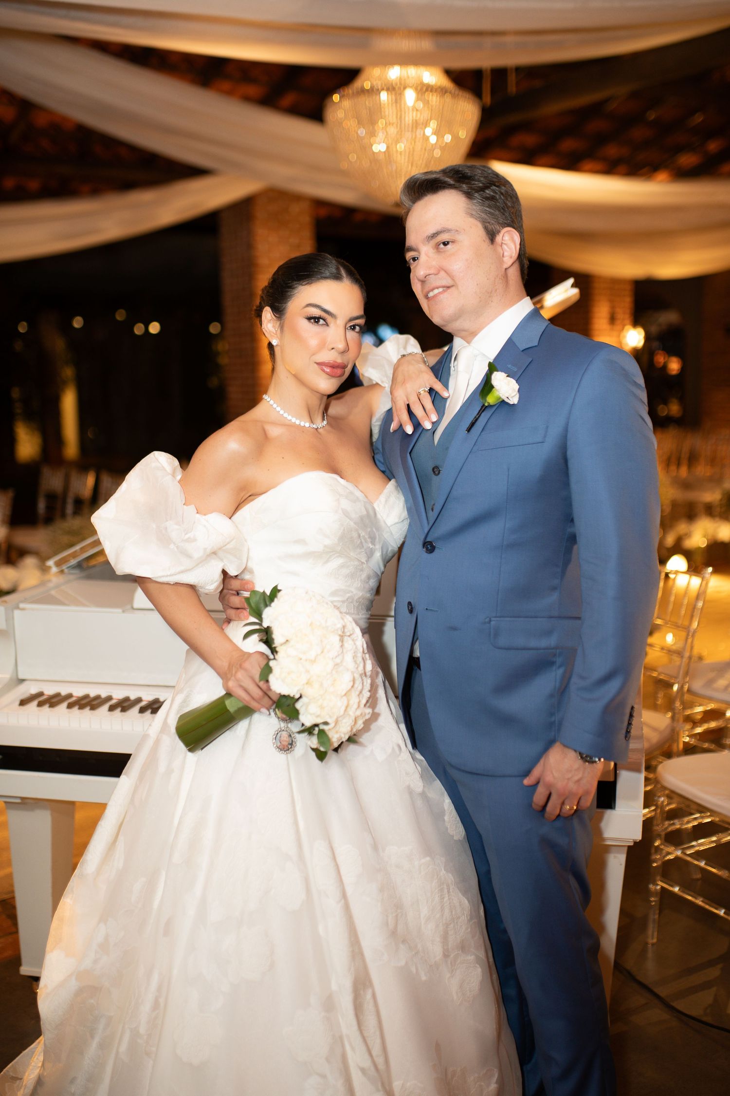Elegant bride in white off-shoulder gown holding bouquet poses with groom in blue suit at wedding reception.