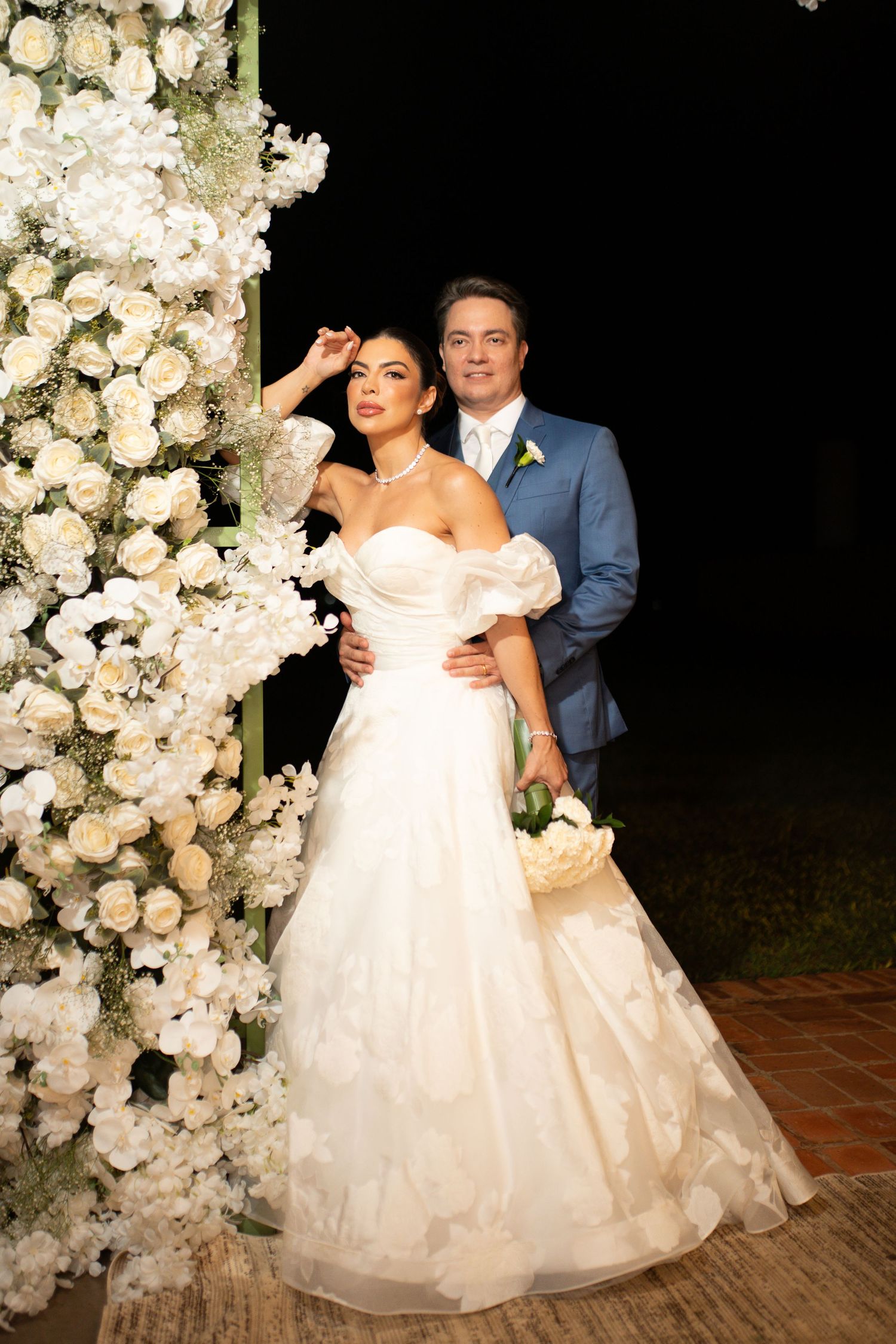 Bride in white off-shoulder gown and groom in blue suit posing by white floral arch at elegant outdoor wedding.