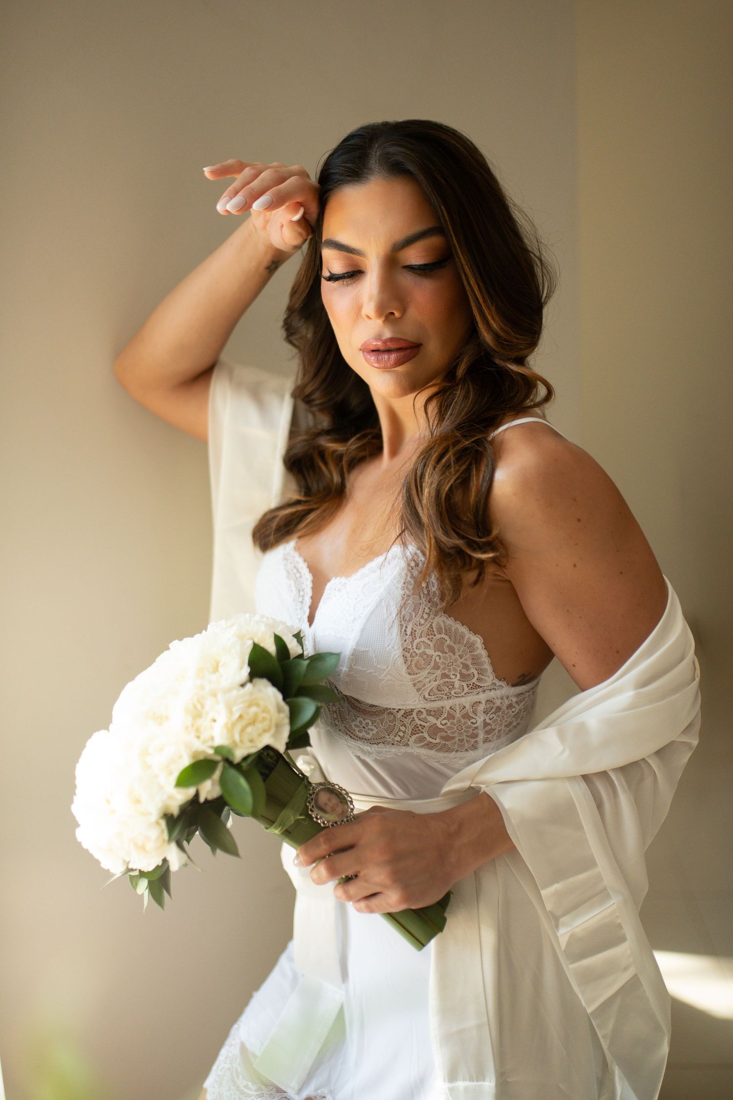 Bride in white lace dress holding bouquet of white roses, posing elegantly with hand near hair.