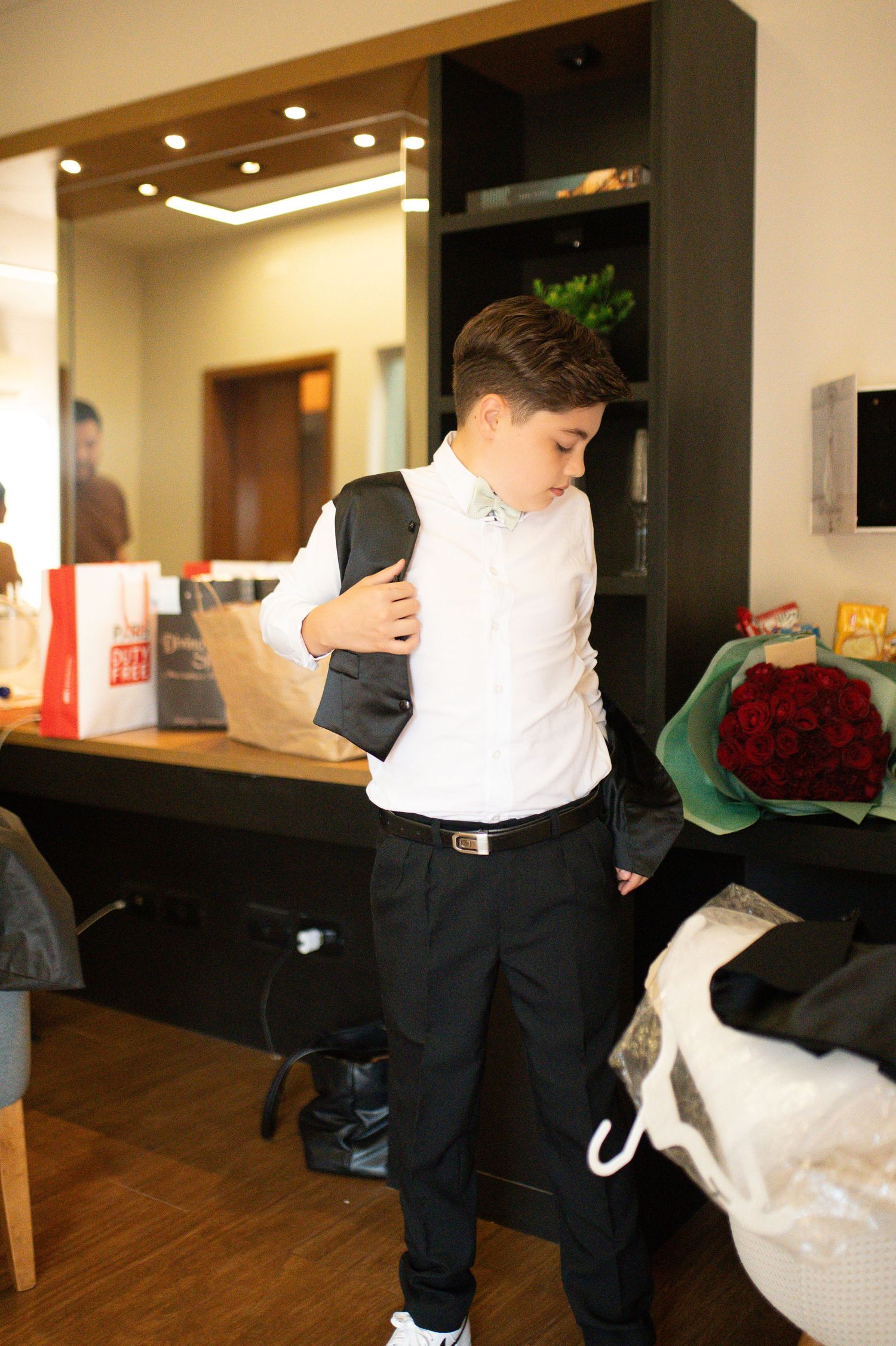 Young boy in white shirt and black pants putting on a suit jacket indoors near a bouquet of red roses.