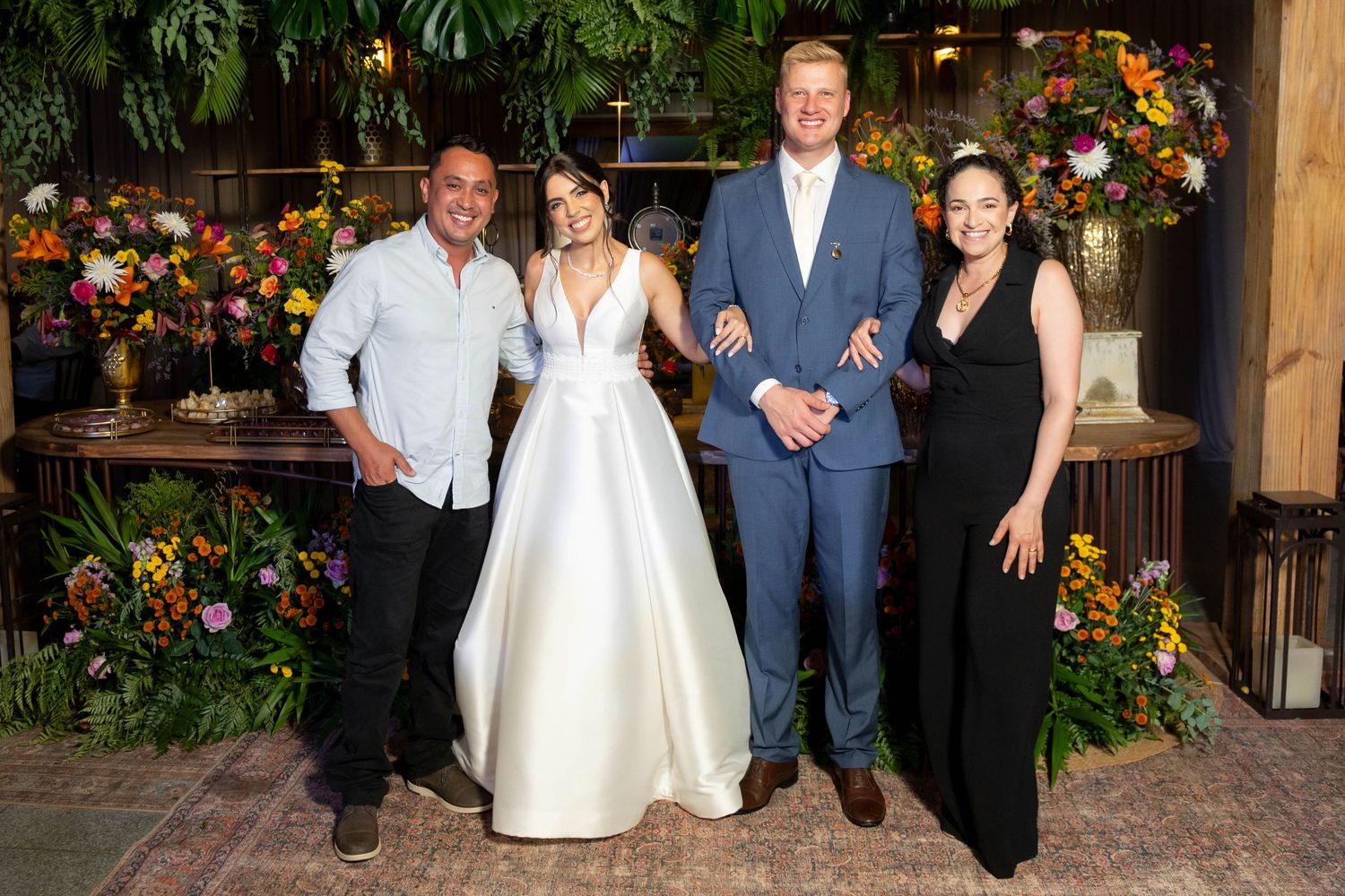 Four people pose together in a floral-decorated venue, one woman wearing a white wedding gown and a man in a blue suit.