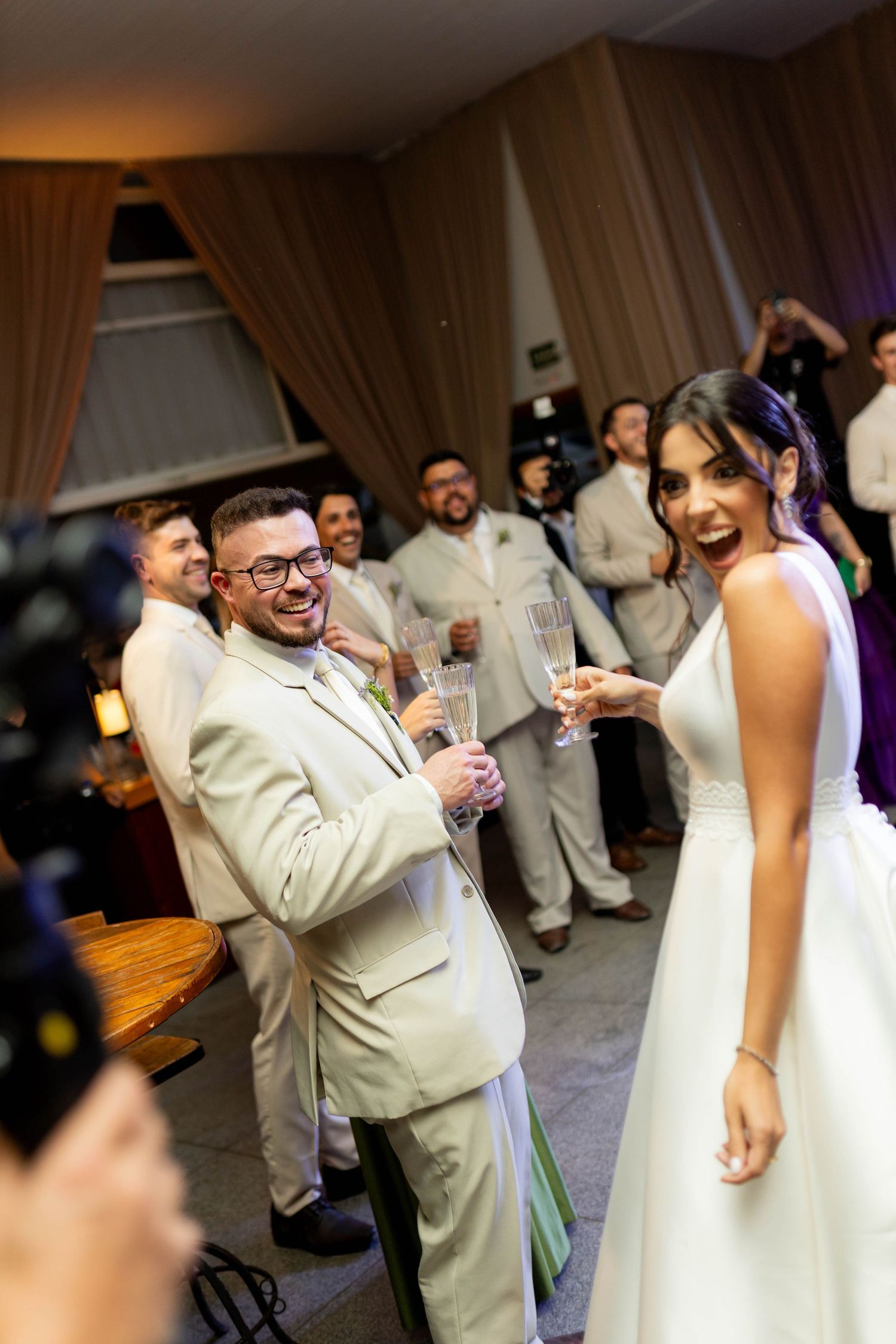 Joyful bride and groom toasting with champagne glasses at their wedding reception surrounded by guests.
