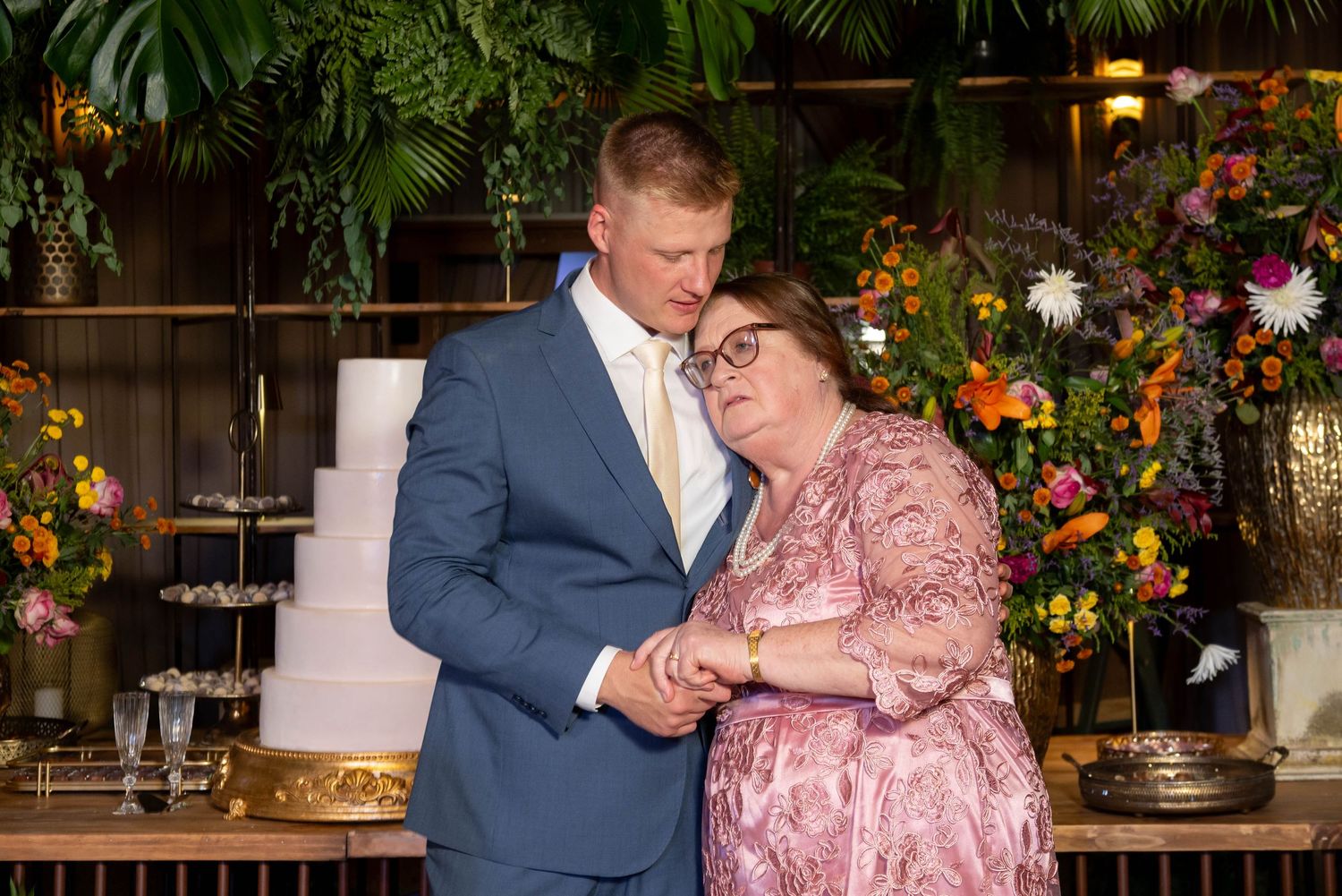 Couple sharing a tender moment at a wedding reception with floral backdrop and white tiered cake.