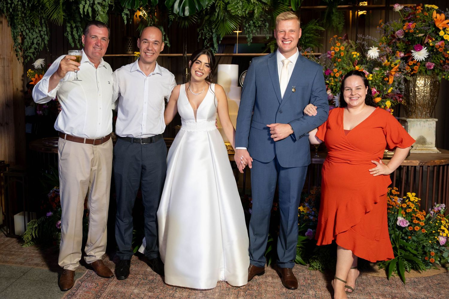 Wedding party of five posing and smiling at elegant floral reception venue, bride in white gown with groom in blue suit.