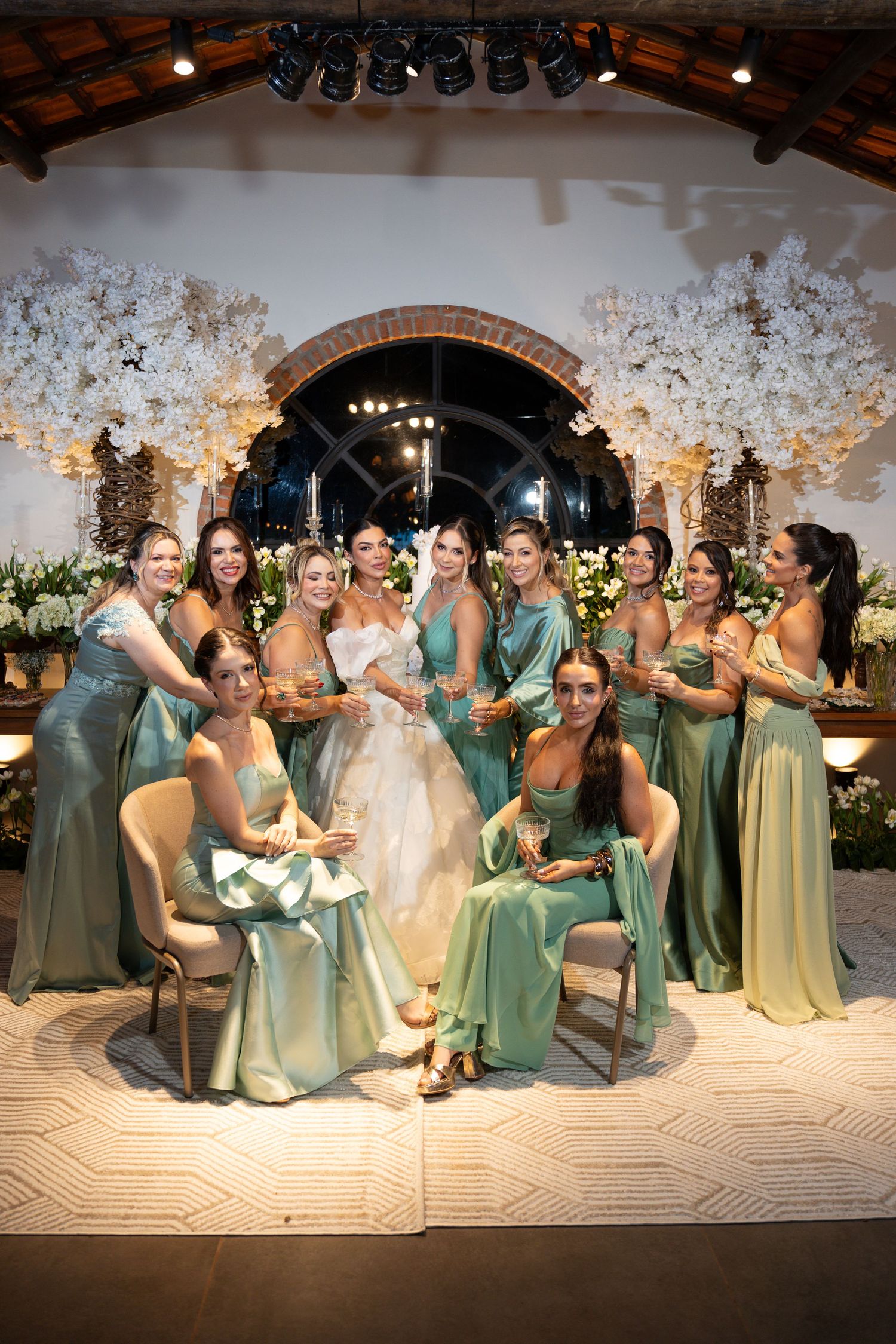Bride in white gown poses with bridesmaids in sage green satin dresses at elegant floral wedding reception.
