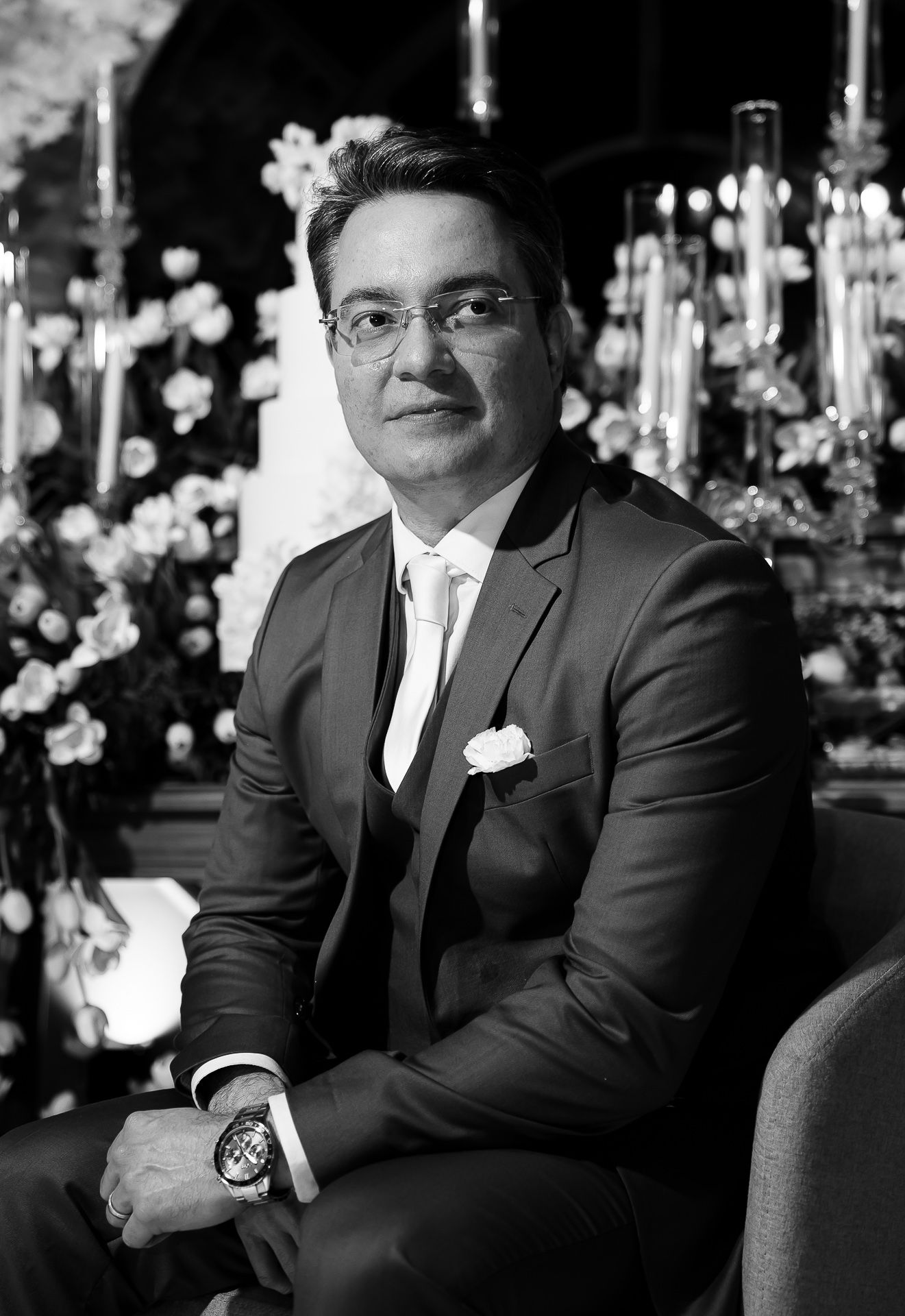 Black and white portrait of an elegant man in a suit with tie and glasses at a formal floral event.