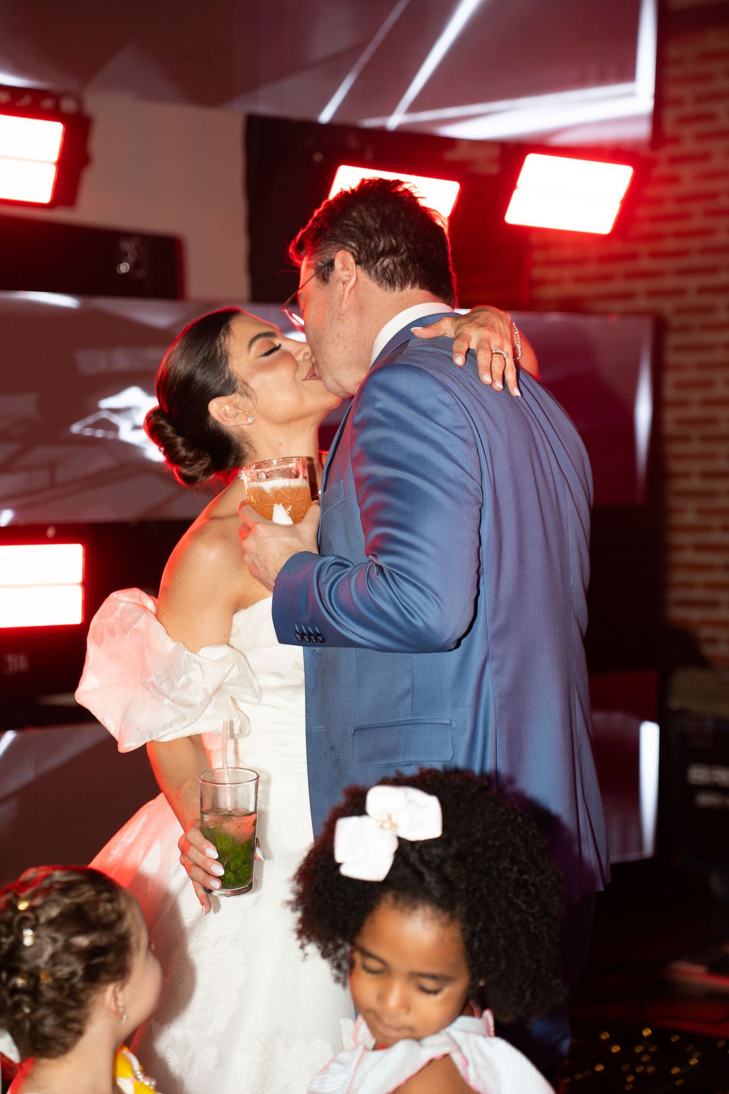 Bride and groom share a kiss at their wedding reception under colorful stage lights, with a young flower girl nearby.