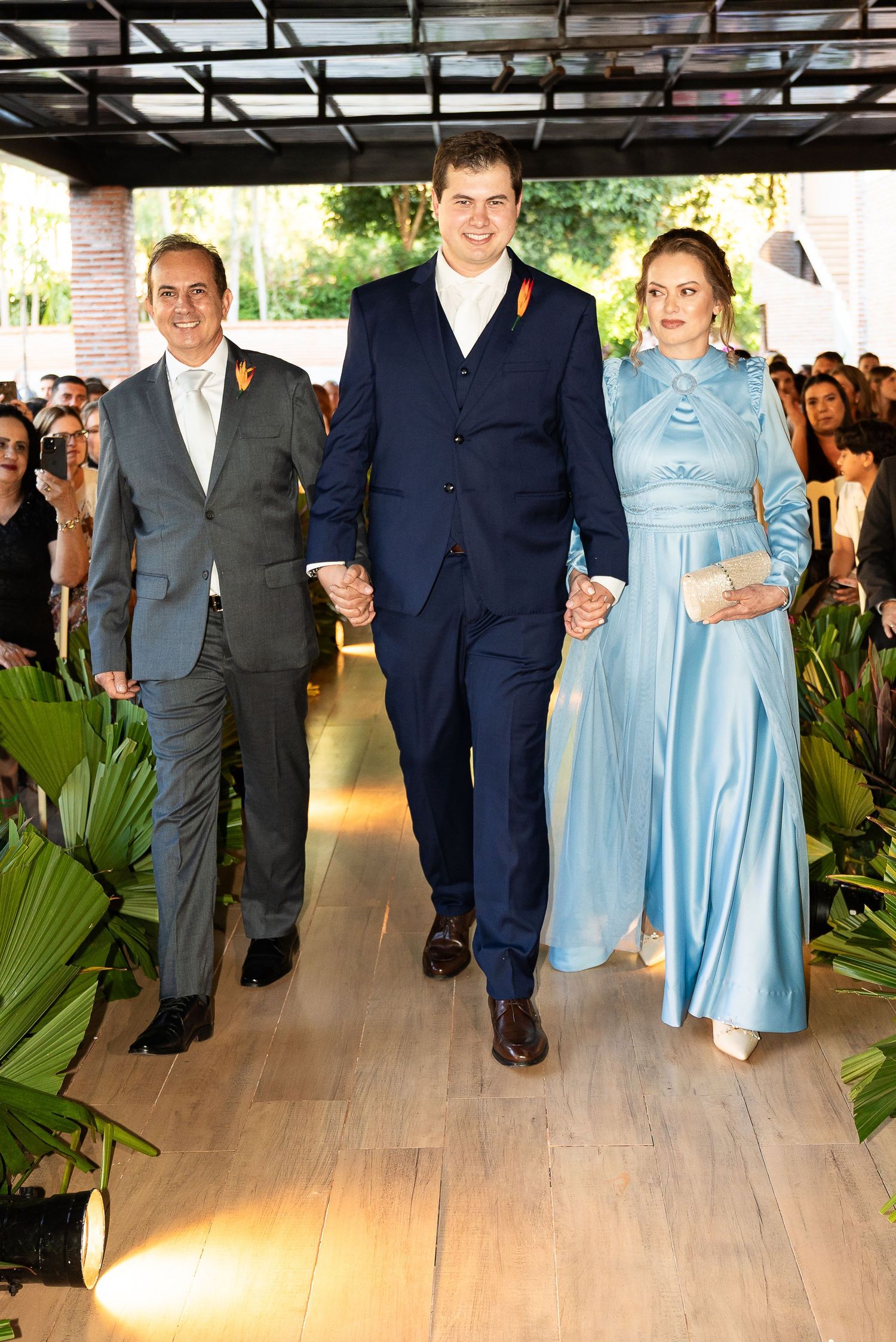 Groom in navy suit walks down wedding aisle with parents, father in gray suit and mother in light blue gown.