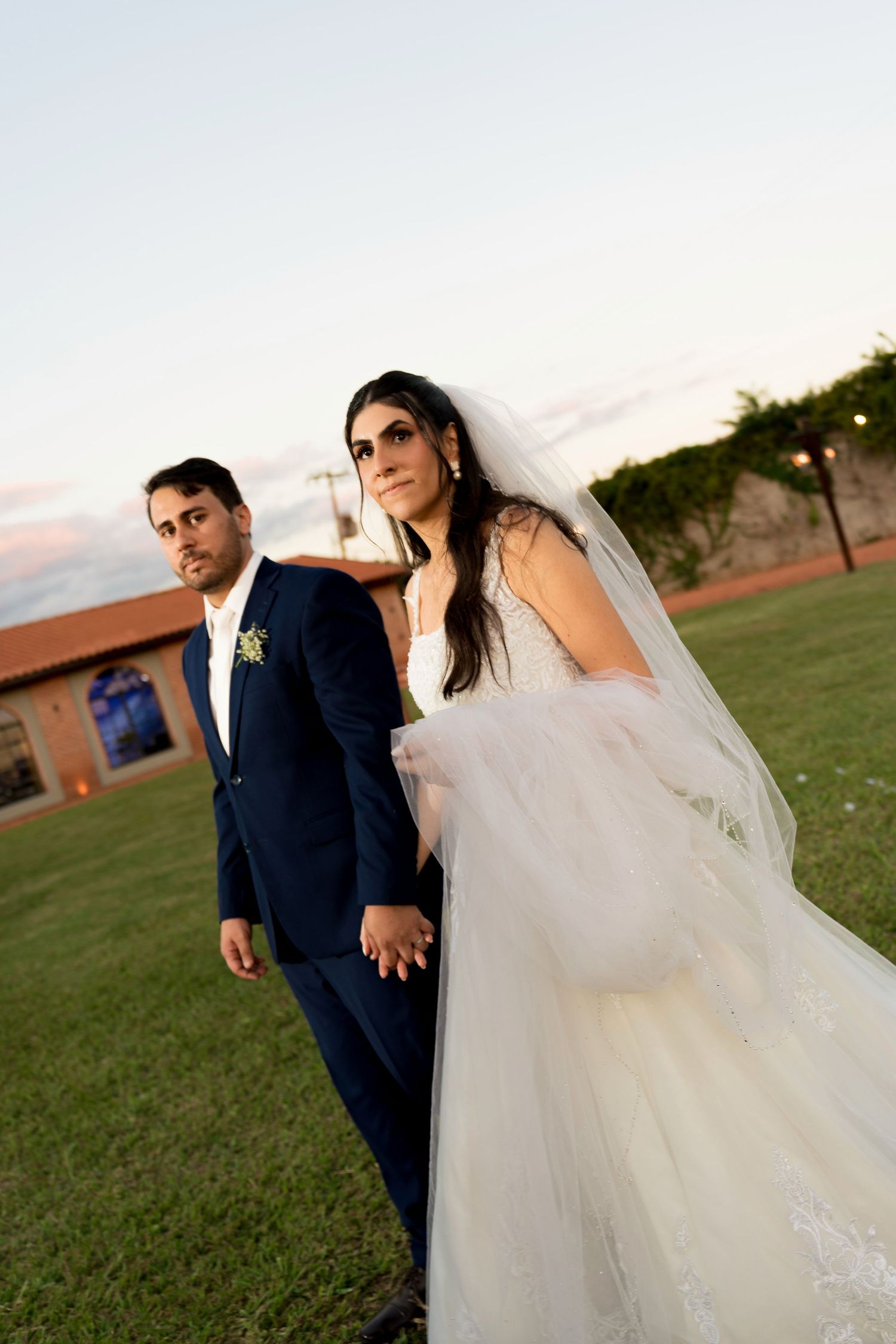 Bride in white gown and veil holds hands with groom in navy suit outdoors at dusk near a brick building.