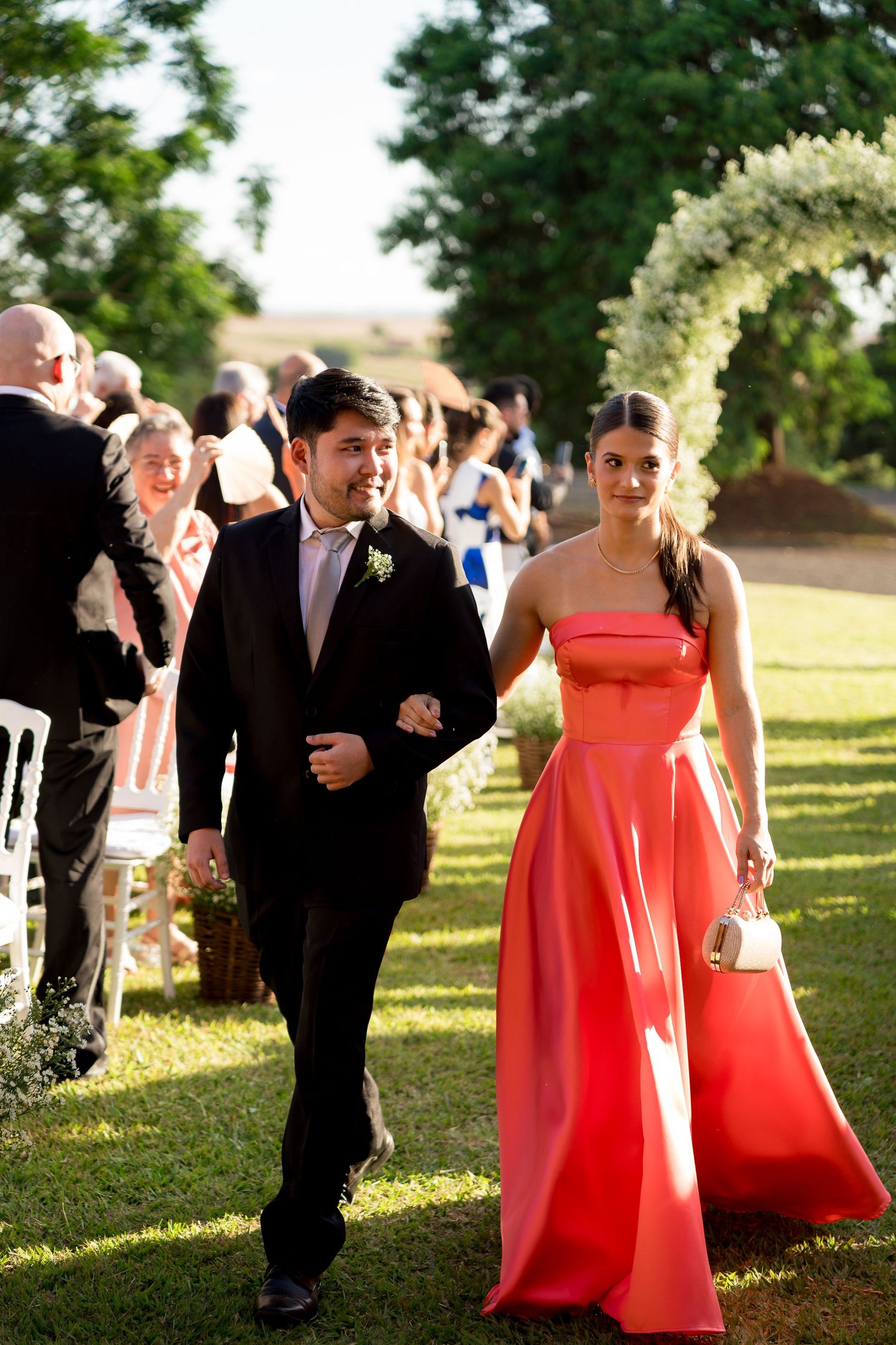 A groomsman in a black suit escorts a bridesmaid in a coral strapless gown down the aisle at an outdoor wedding.