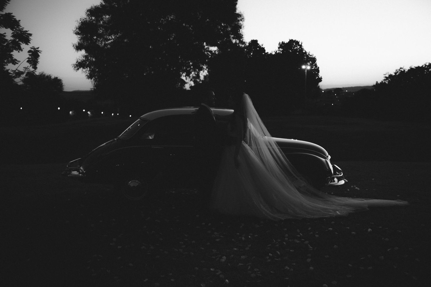 Black and white wedding photo of a bride in a flowing veil beside a vintage car at dusk.