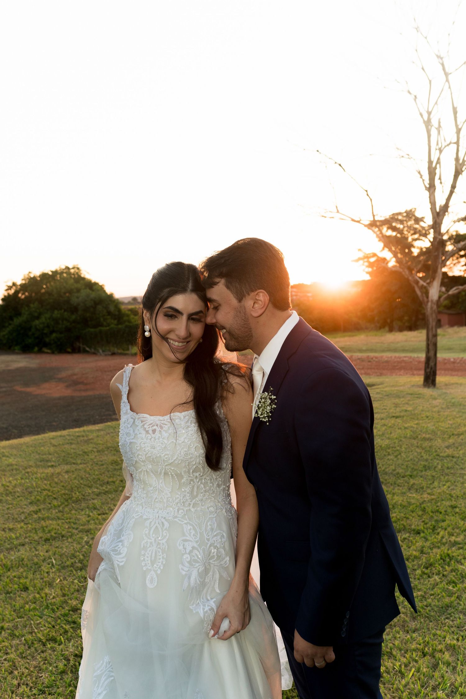 Bride and groom share a romantic sunset moment outdoors, with golden light glowing behind them on a green lawn.