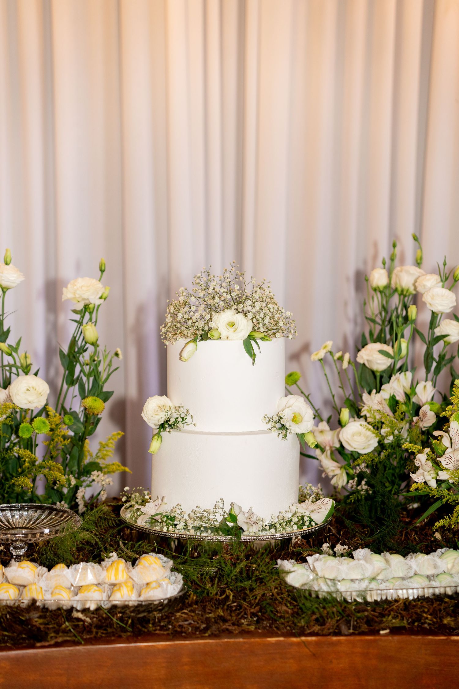 Elegant two-tier white wedding cake adorned with baby's breath and roses, surrounded by lush white floral arrangements.