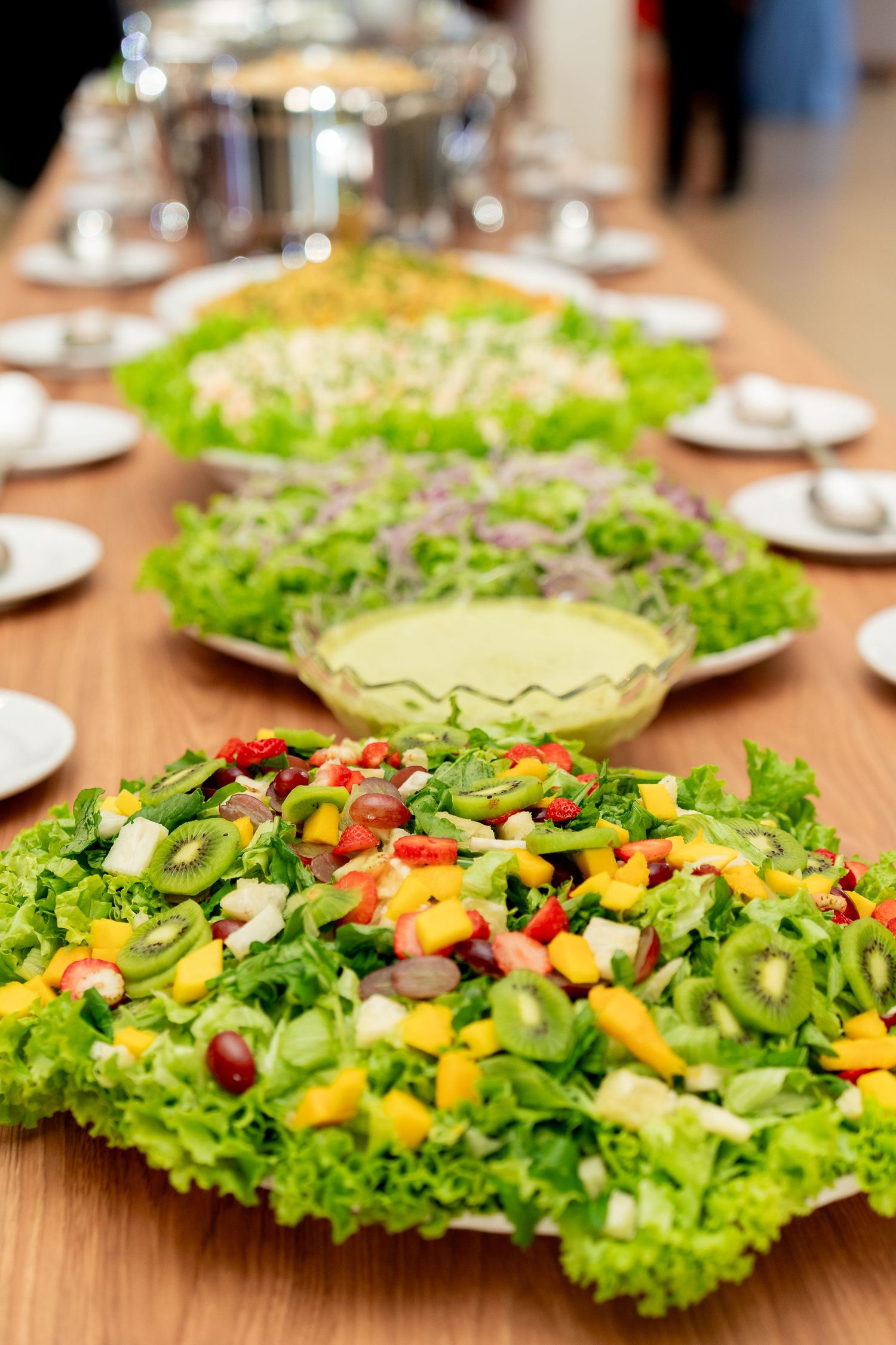 Colorful fresh fruit and vegetable salads displayed on a wooden buffet table with dressing bowls and white plates.
