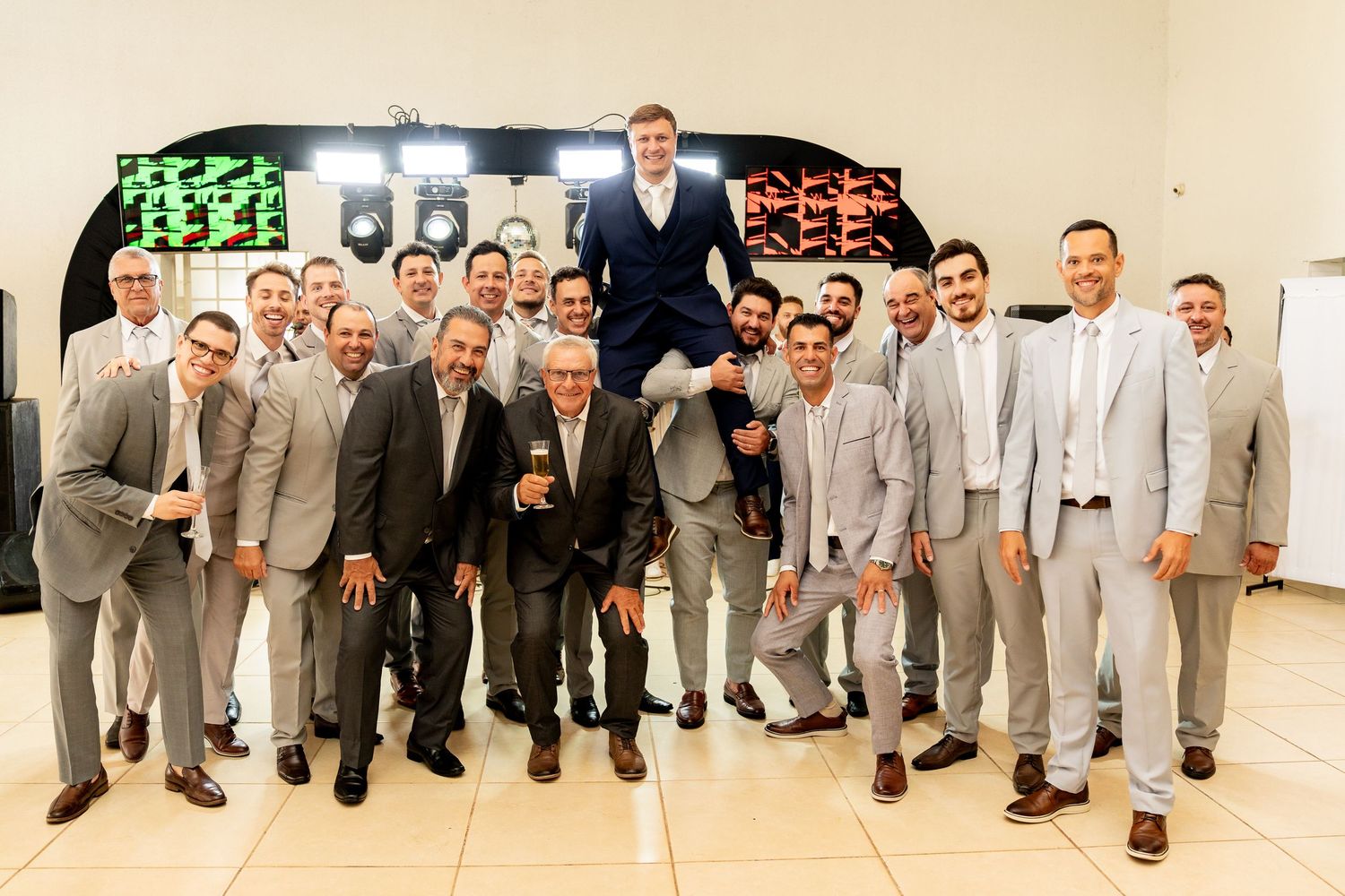 Large group of men in matching gray suits posing together at a formal event, with one man lifted up in celebration.