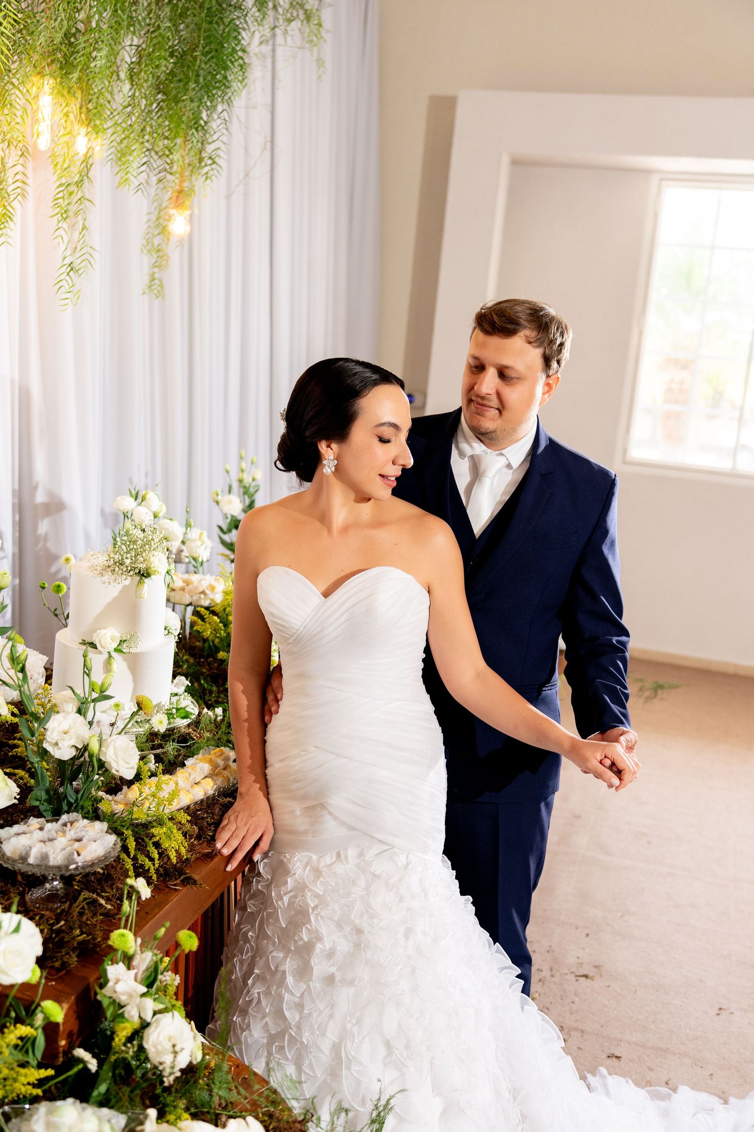 Bride in strapless mermaid gown and groom in navy suit share a tender moment beside floral arrangements at their wedding.