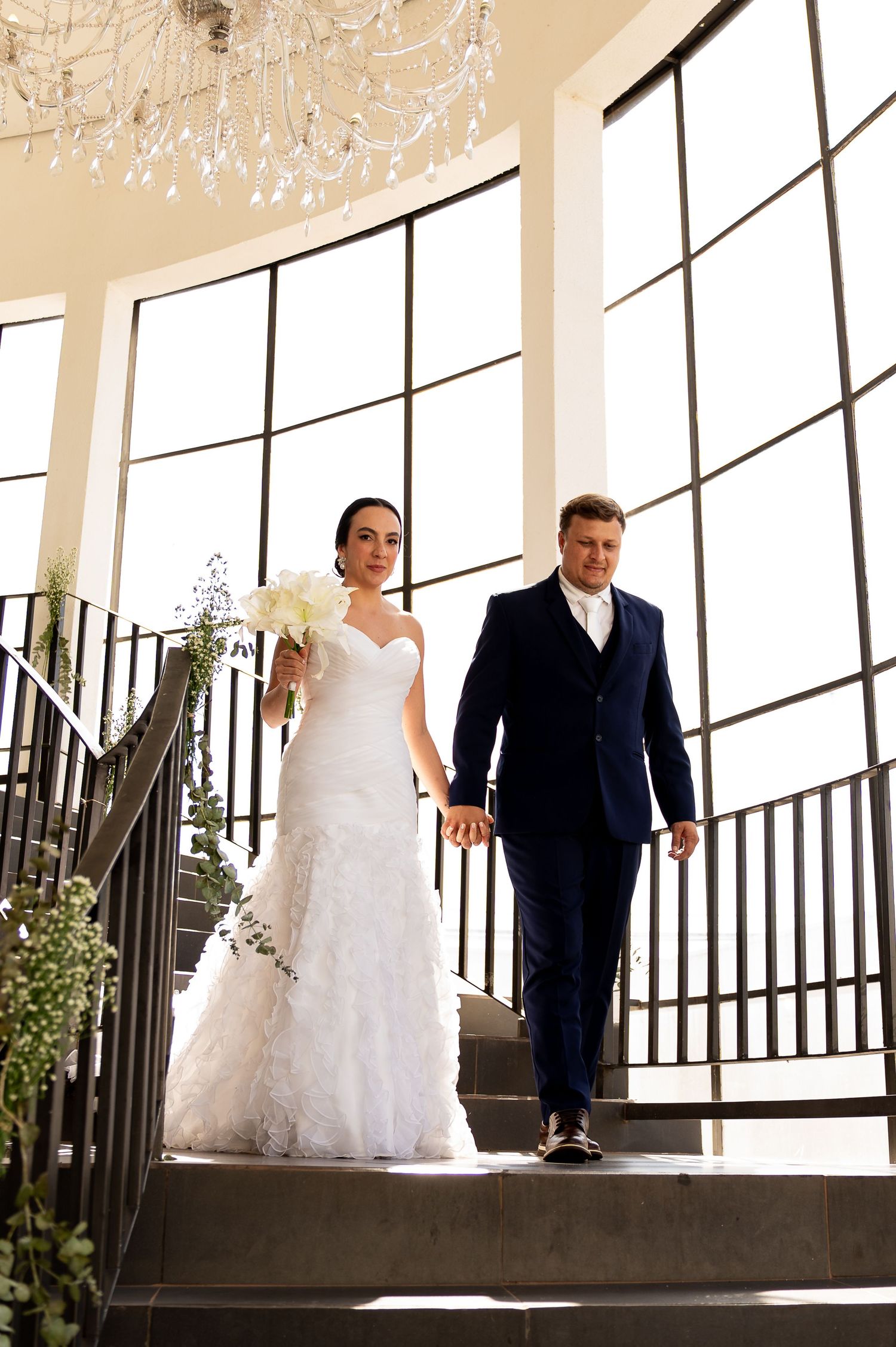 Bride in white mermaid gown and groom in navy suit descend elegant staircase with chandelier backdrop.
