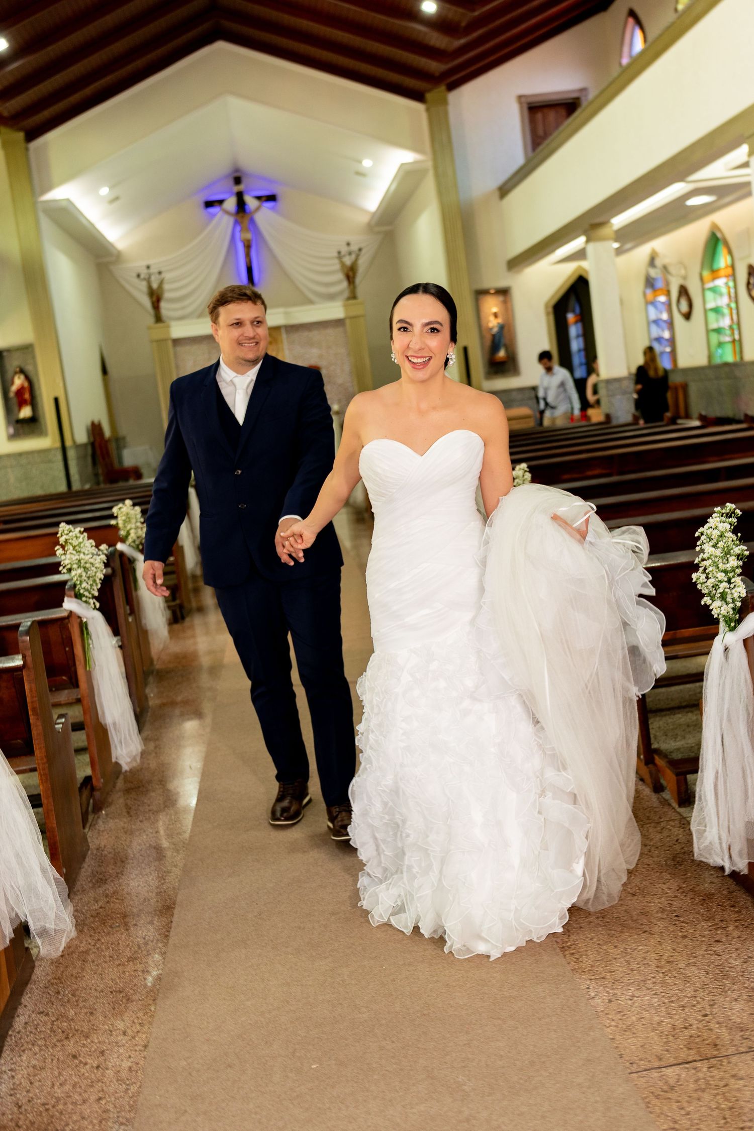 Bride and groom walk hand in hand down church aisle after wedding ceremony, bride in white ruffled gown.