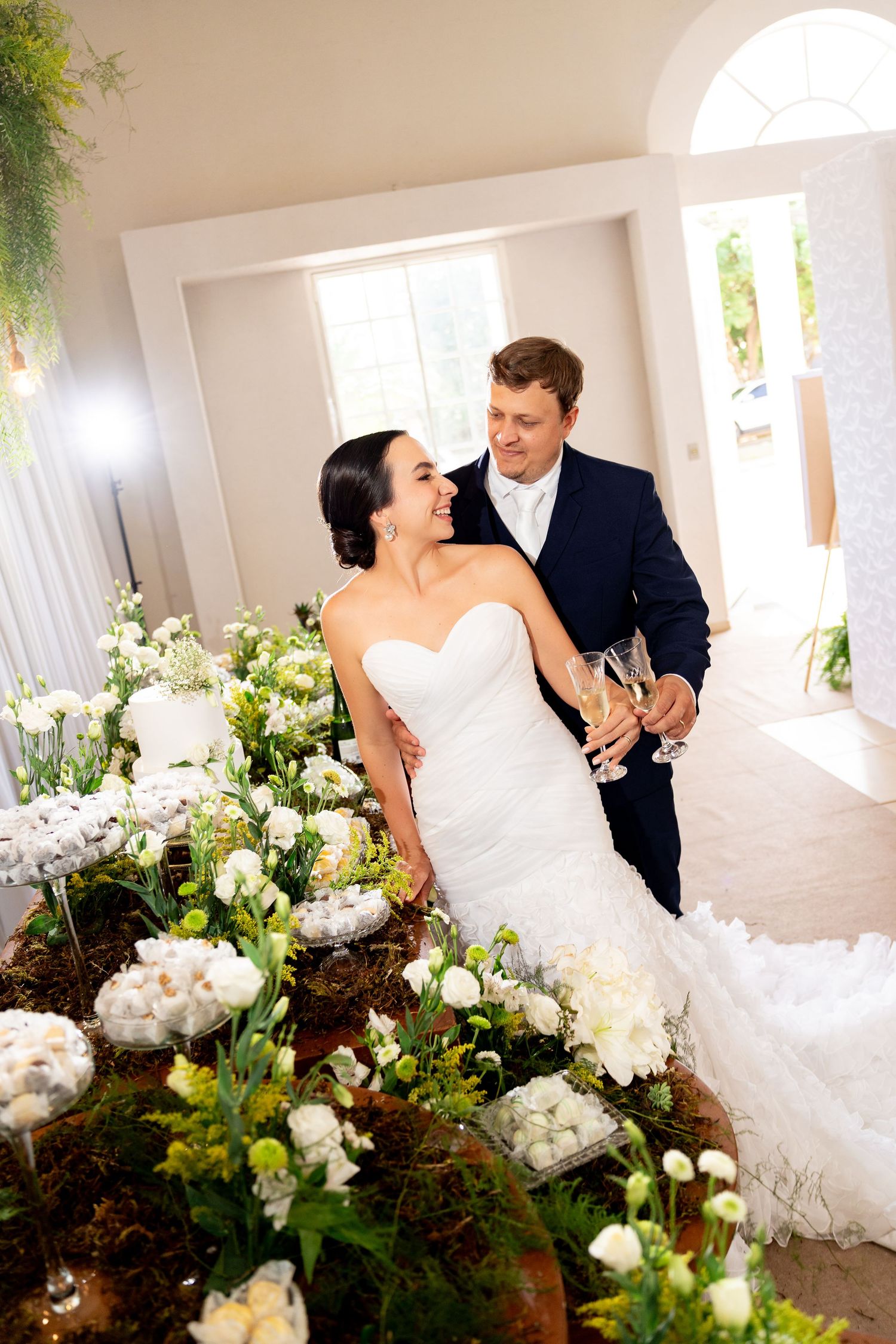 Bride and groom toast with champagne flutes beside elegant white floral arrangements at their wedding reception.