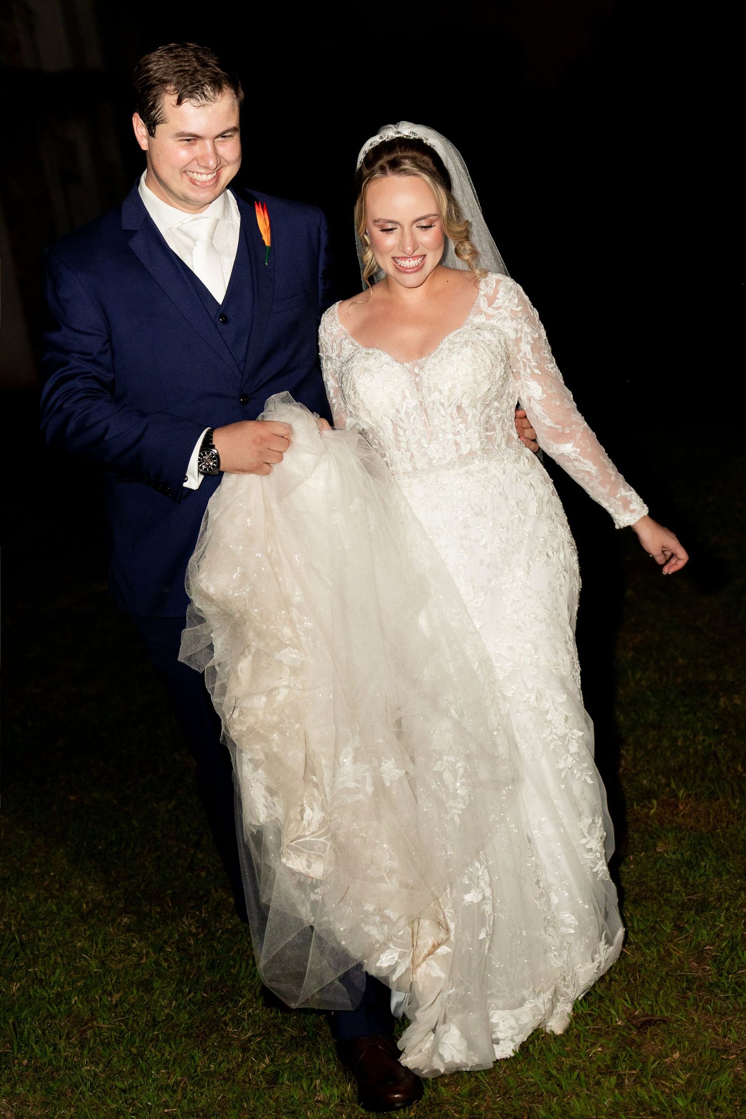 Bride in lace gown and groom in navy suit walk together at night during their wedding celebration.