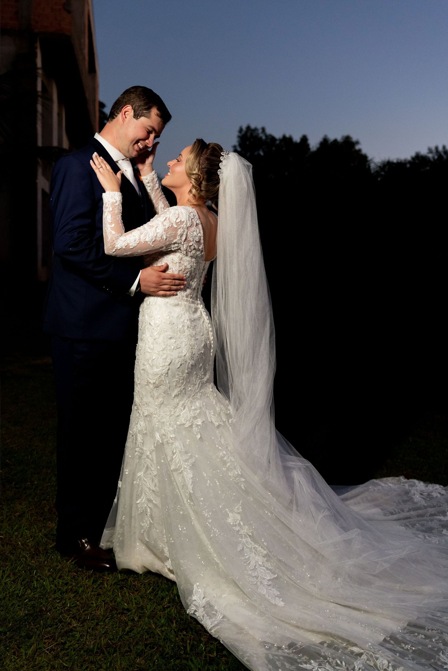 Bride in lace mermaid gown with veil embraces groom in navy suit at romantic outdoor wedding ceremony at dusk.