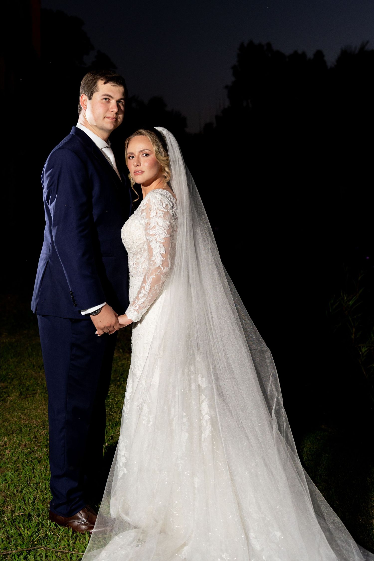 Bride in lace gown with veil and groom in navy suit hold hands posing outdoors at night.