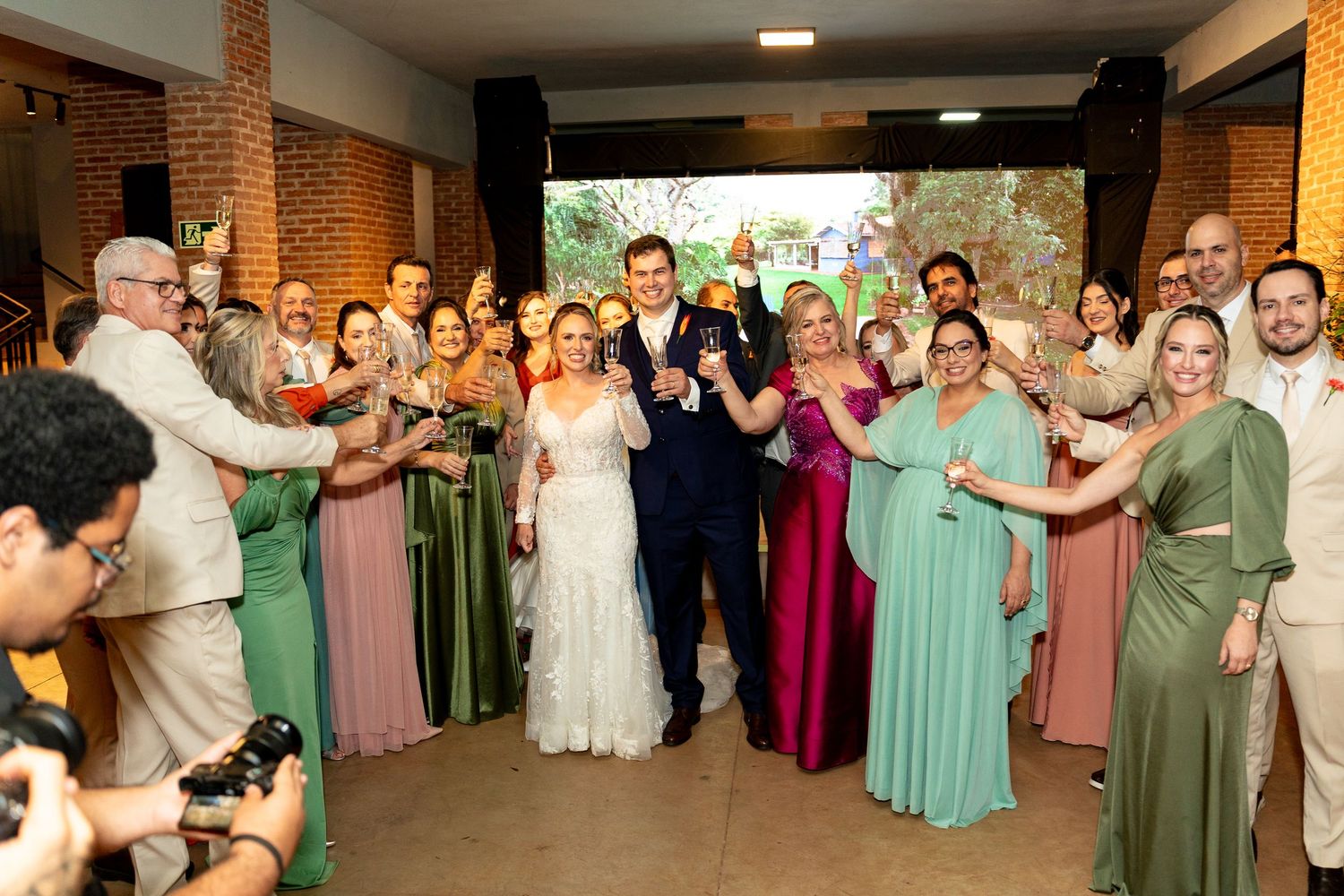 Bride and groom celebrate with wedding party and guests, bridesmaids in green and pink dresses, raising glasses in toast.