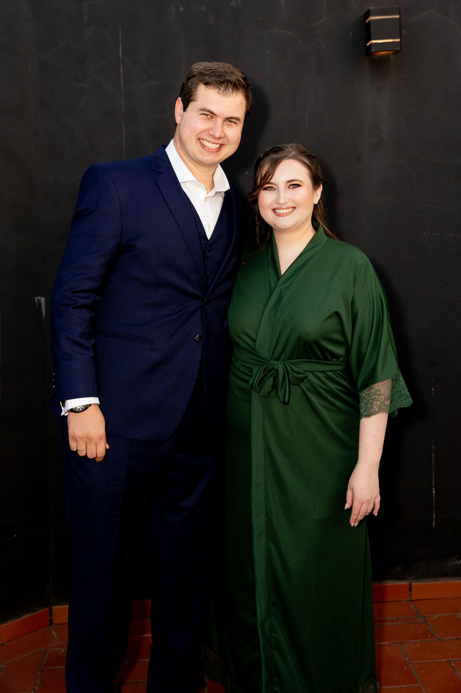 A smiling couple poses together, man in navy suit and woman in elegant green satin dress, against a dark wall.