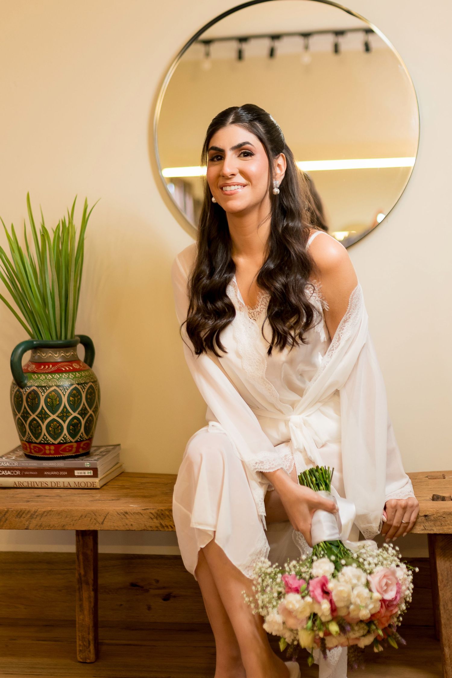 Smiling bride in white robe holding pink floral bouquet seated on wooden bench before round mirror.