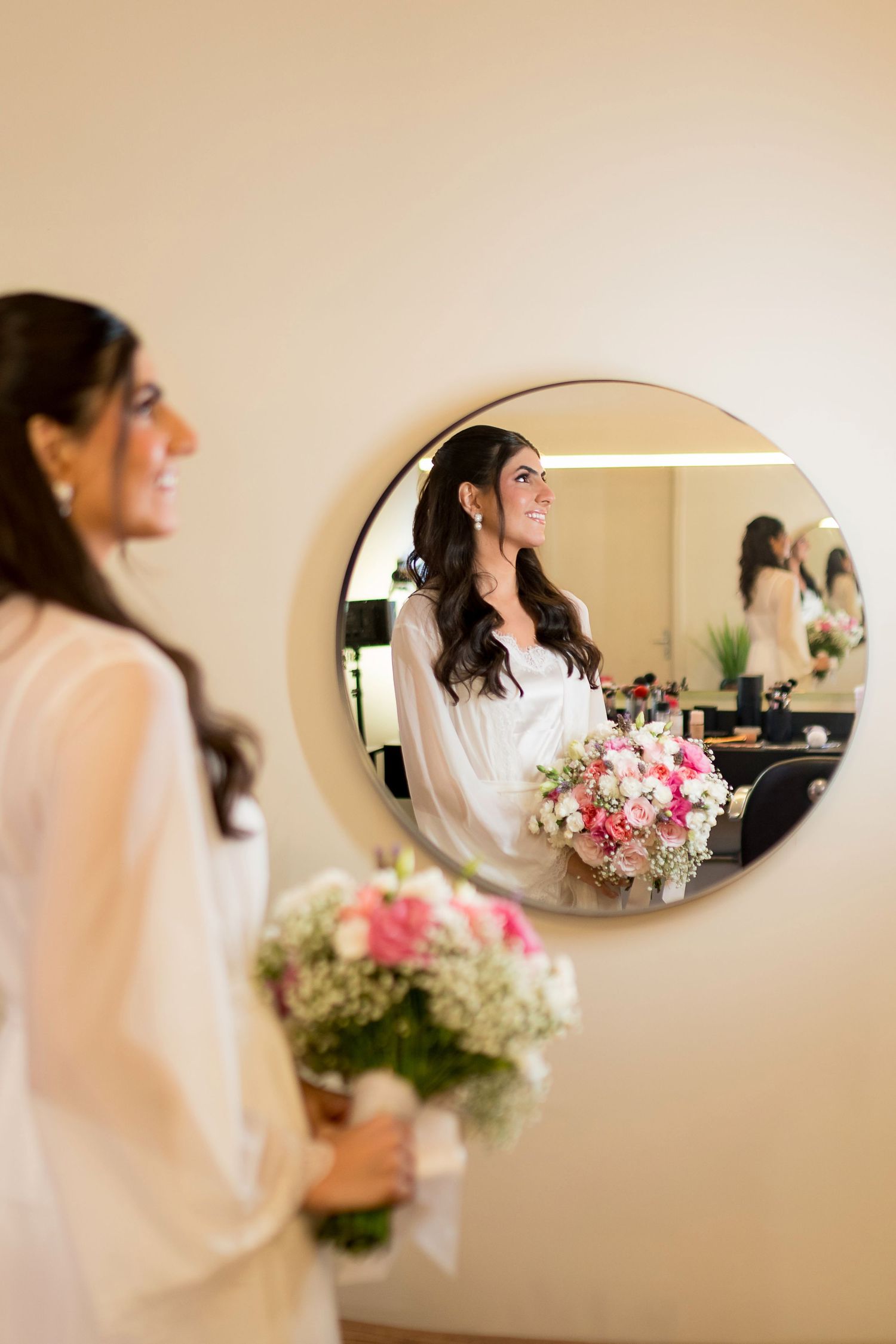 Bride in white robe holds pink bouquet, smiling at her reflection in a round mirror during wedding preparations.
