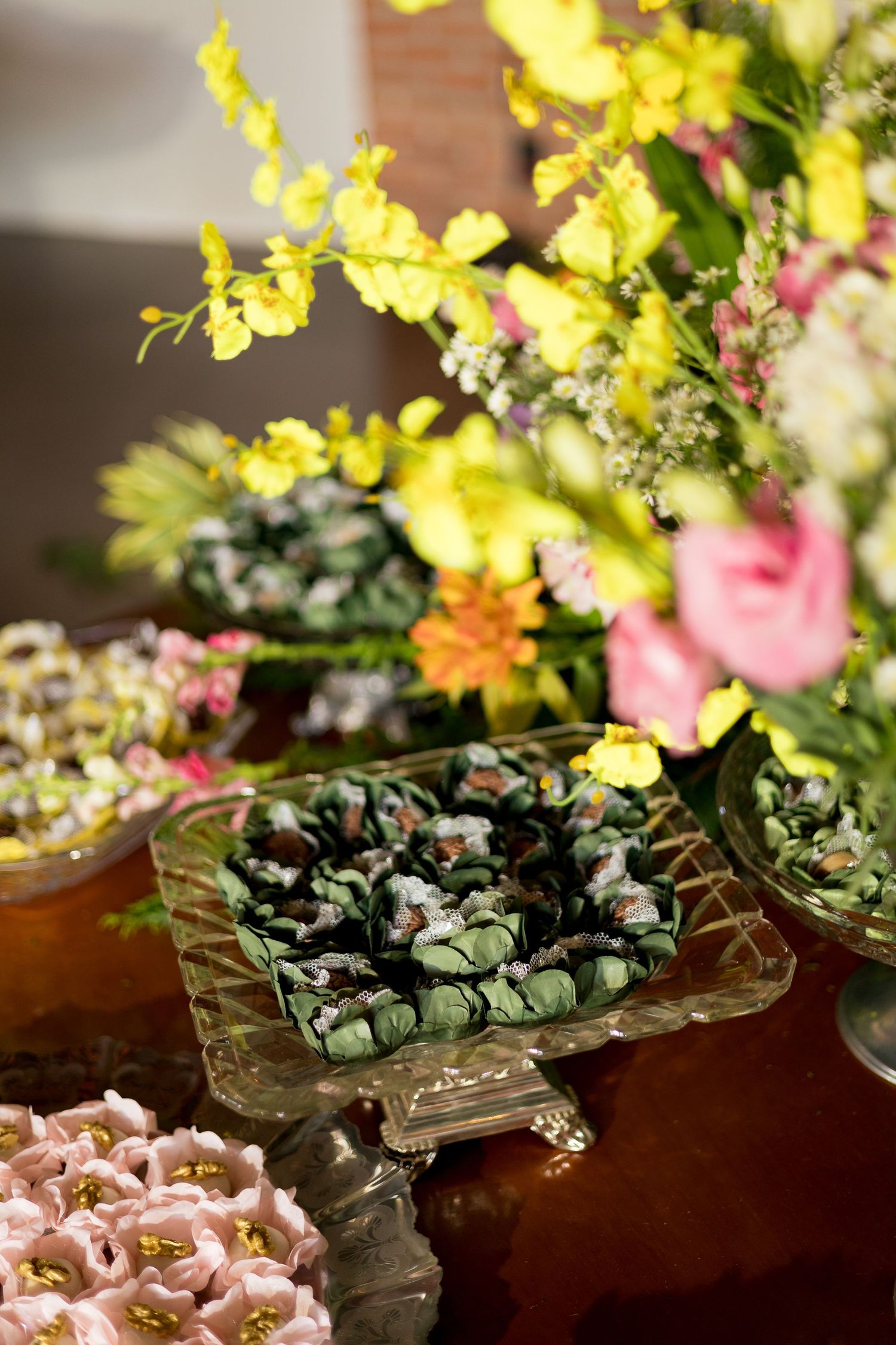 Elegant dessert table with green chocolate truffles on crystal stand, pink sweets, and colorful floral arrangement.