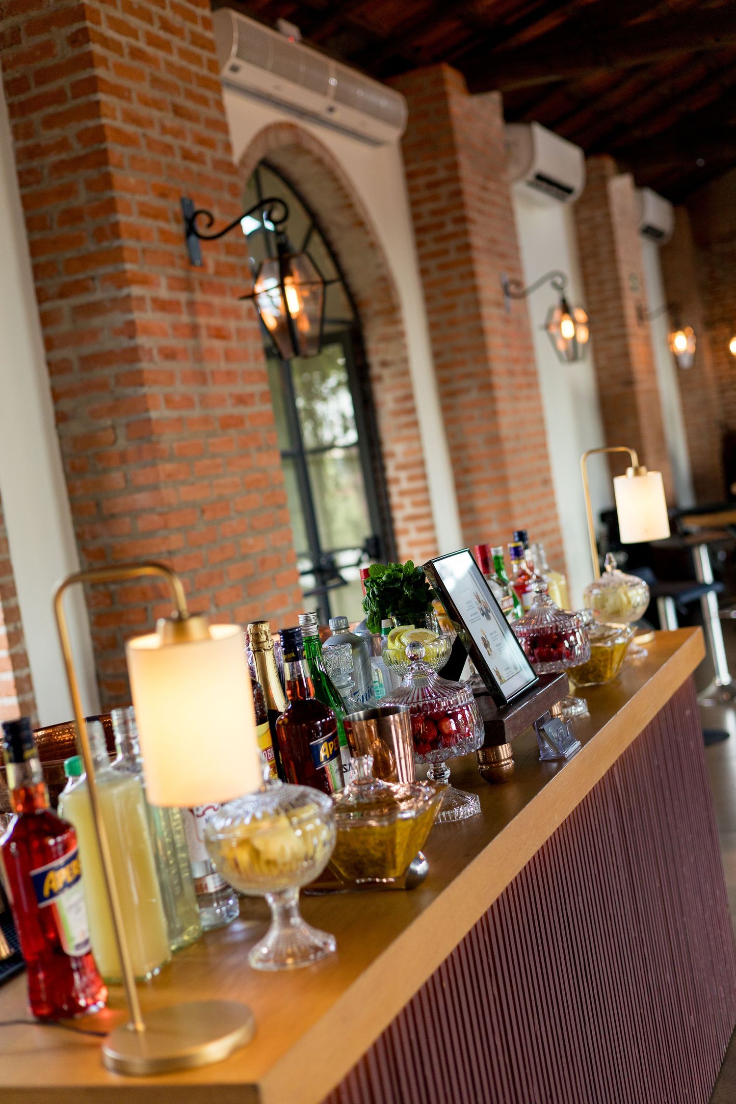 Elegant bar setup with bottles, glowing lamps, and decorative bowls in a rustic brick-walled venue.
