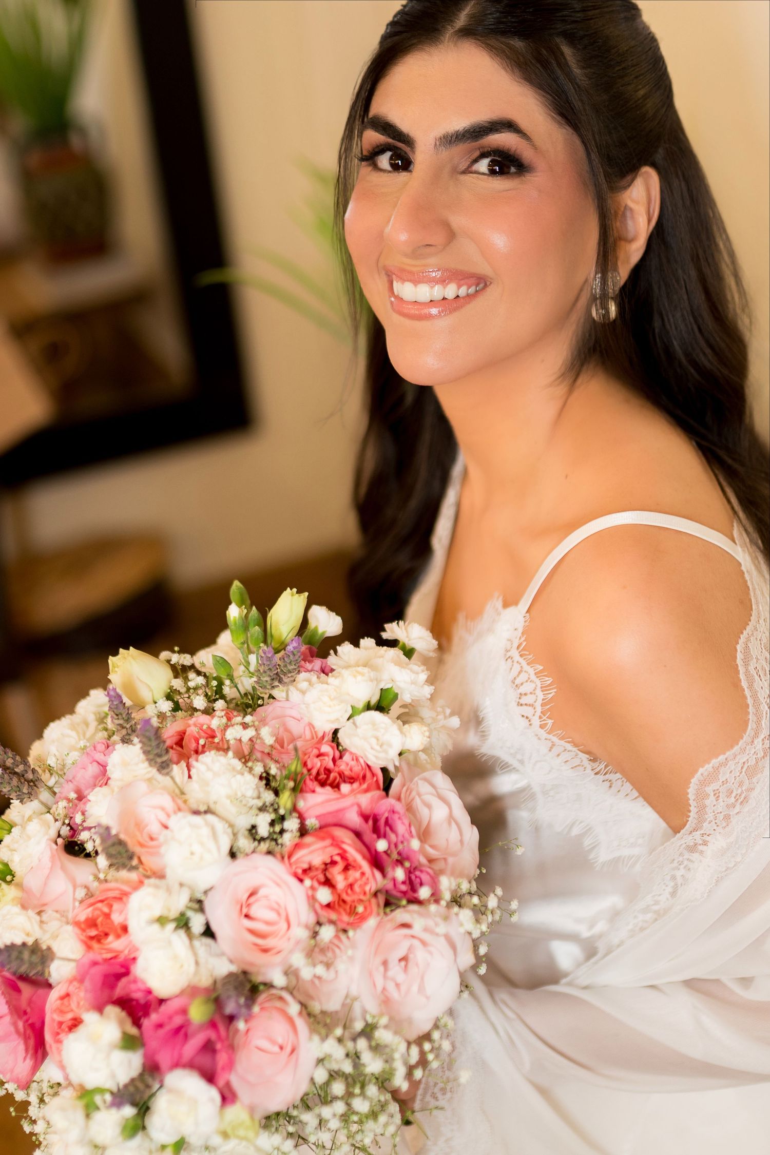Smiling bride in white lace gown holds a lush pink and white floral bouquet with roses and baby's breath.