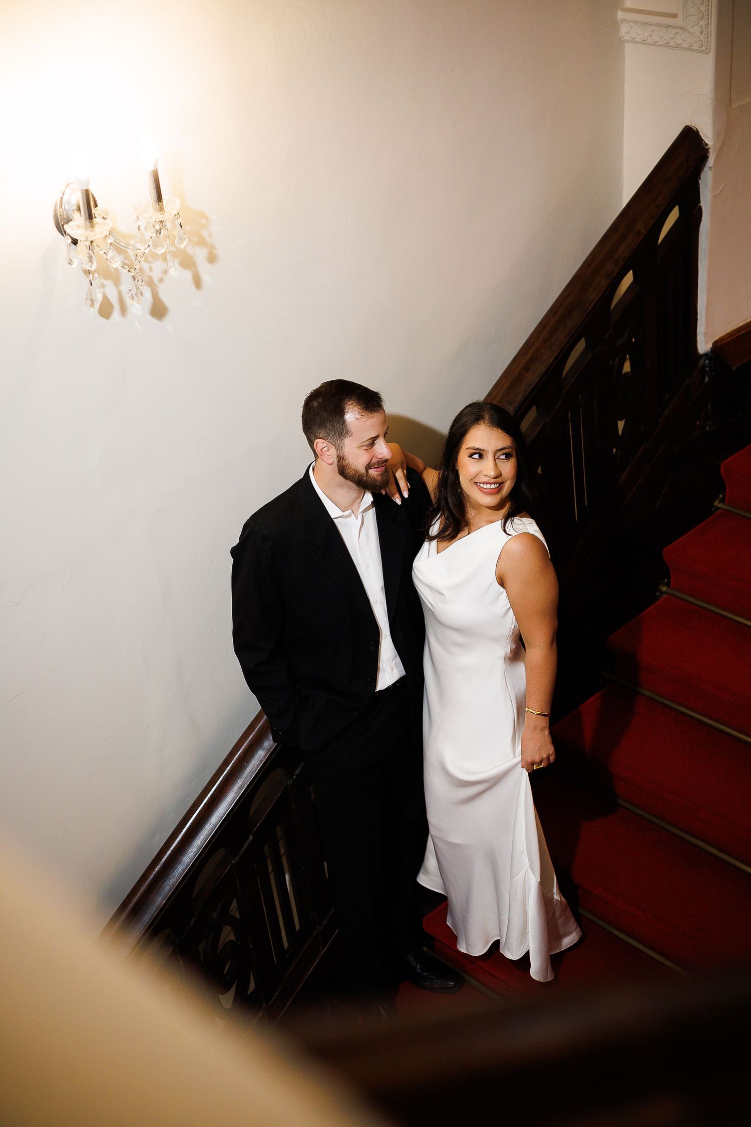 Elegant couple in formal attire posing on a grand staircase with red carpet and wood railings in a classic interior.
