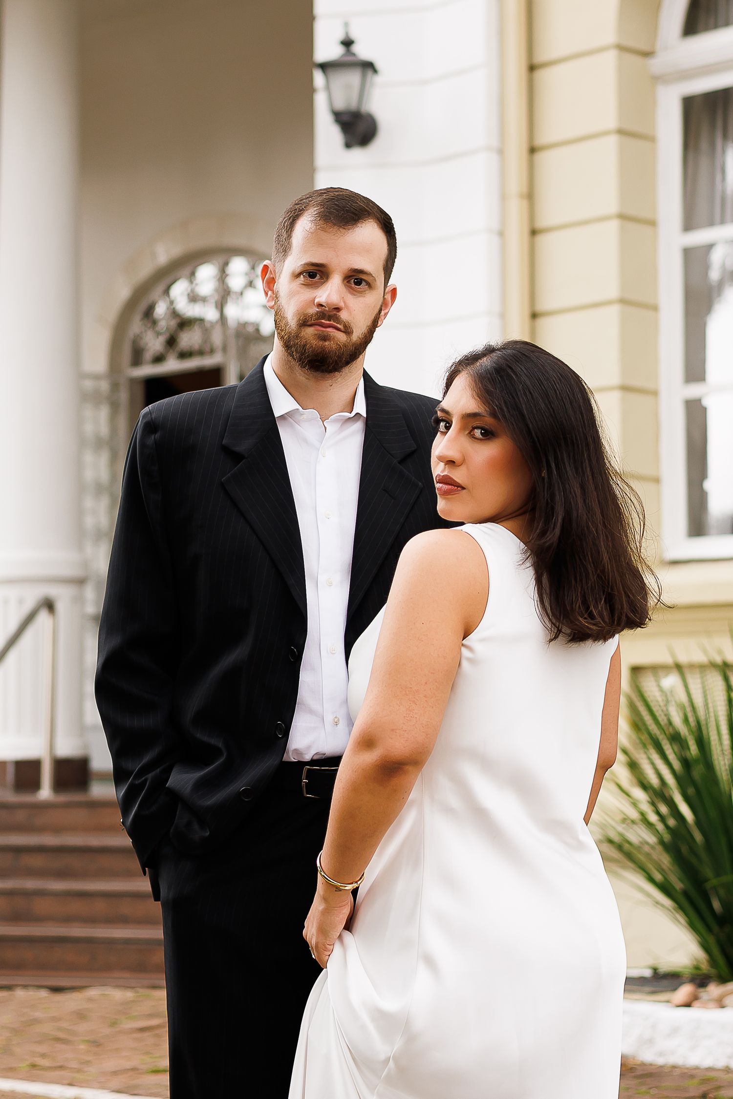 Elegant couple posing outdoors, man in black suit and woman in white dress, standing before a classic building.
