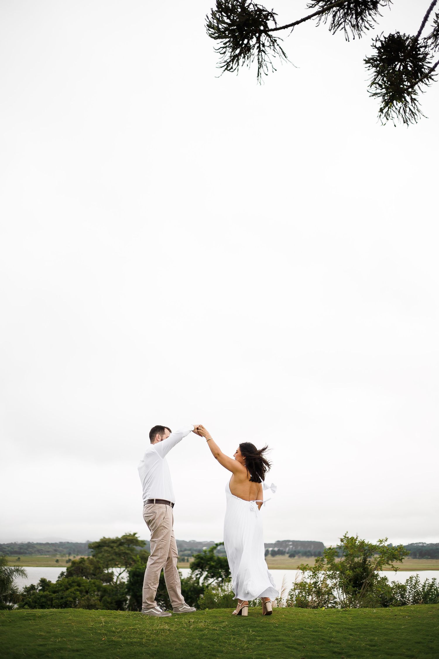 Couple dancing outdoors on green lawn near waterfront, bride in white dress and groom in khaki pants under pine tree.