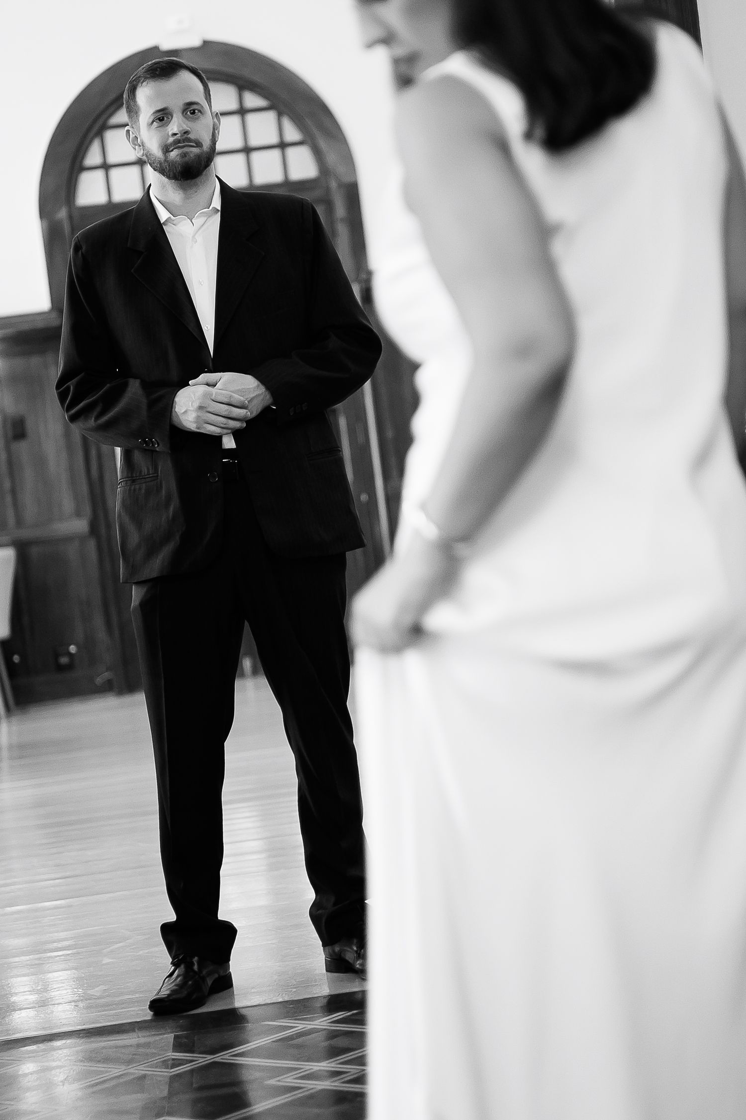 Black and white photo of groom in dark suit smiling at bride in white dress during wedding first look.