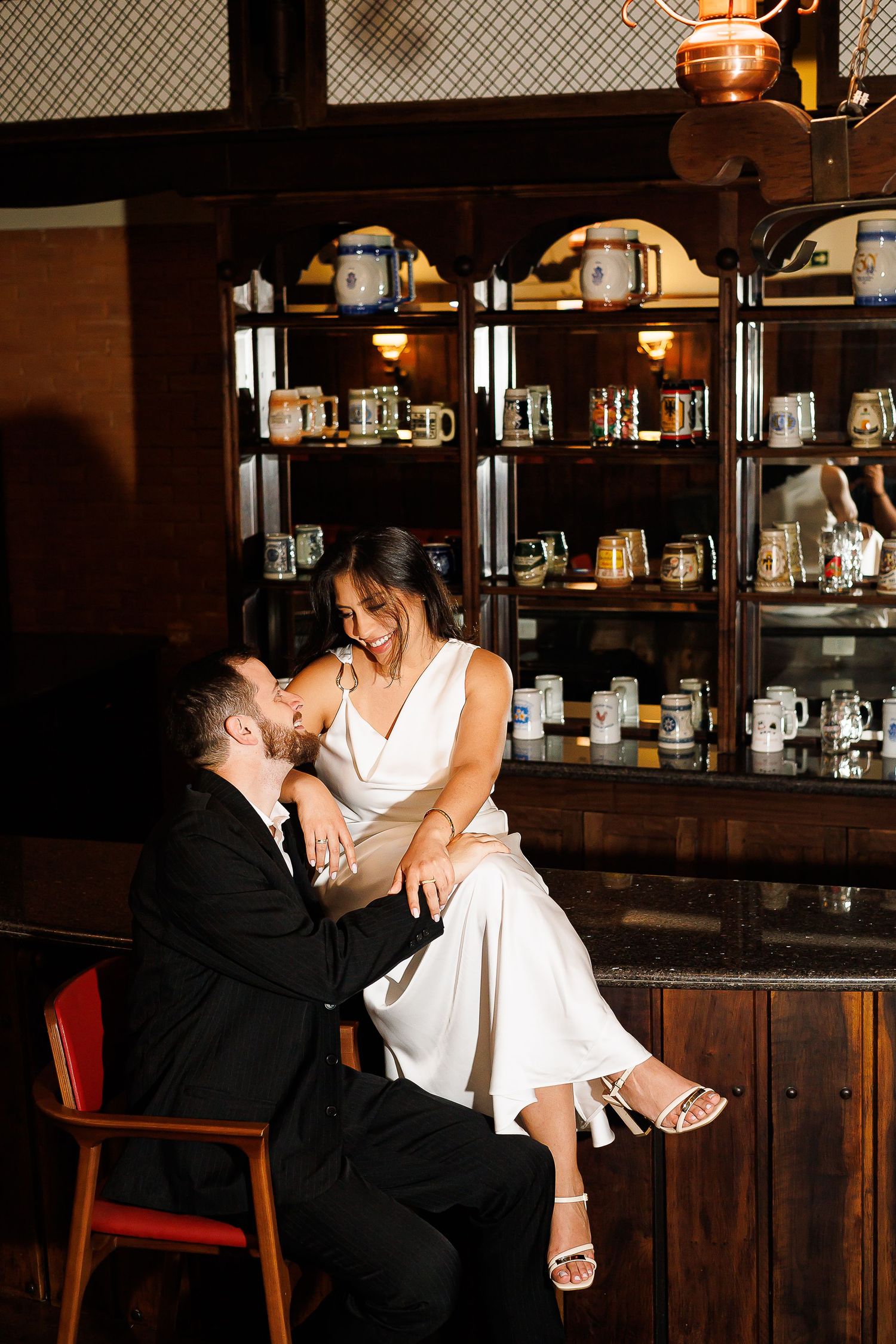 Couple in wedding attire sharing a romantic moment at a vintage bar with shelves of decorative mugs behind them.