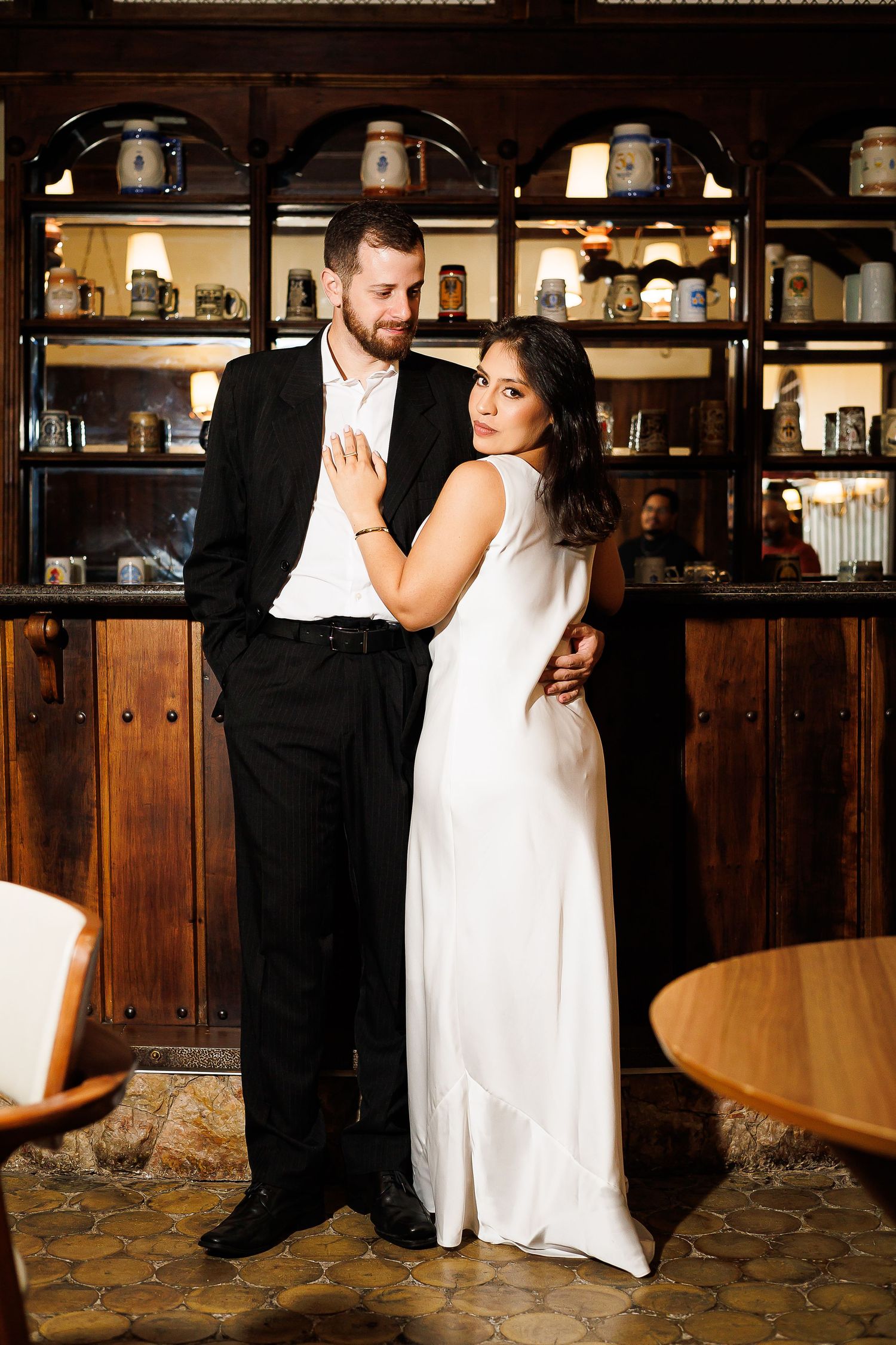 Elegant couple in wedding attire posing in a rustic bar, groom in black suit, bride in white gown.