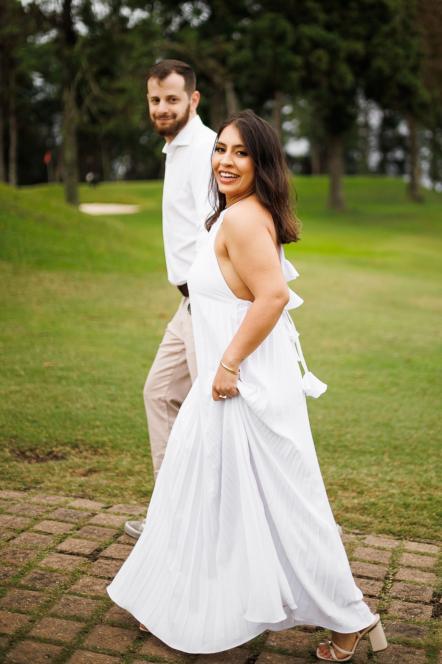 Smiling bride in white halter gown walks with groom in white shirt on brick path at lush green golf course.