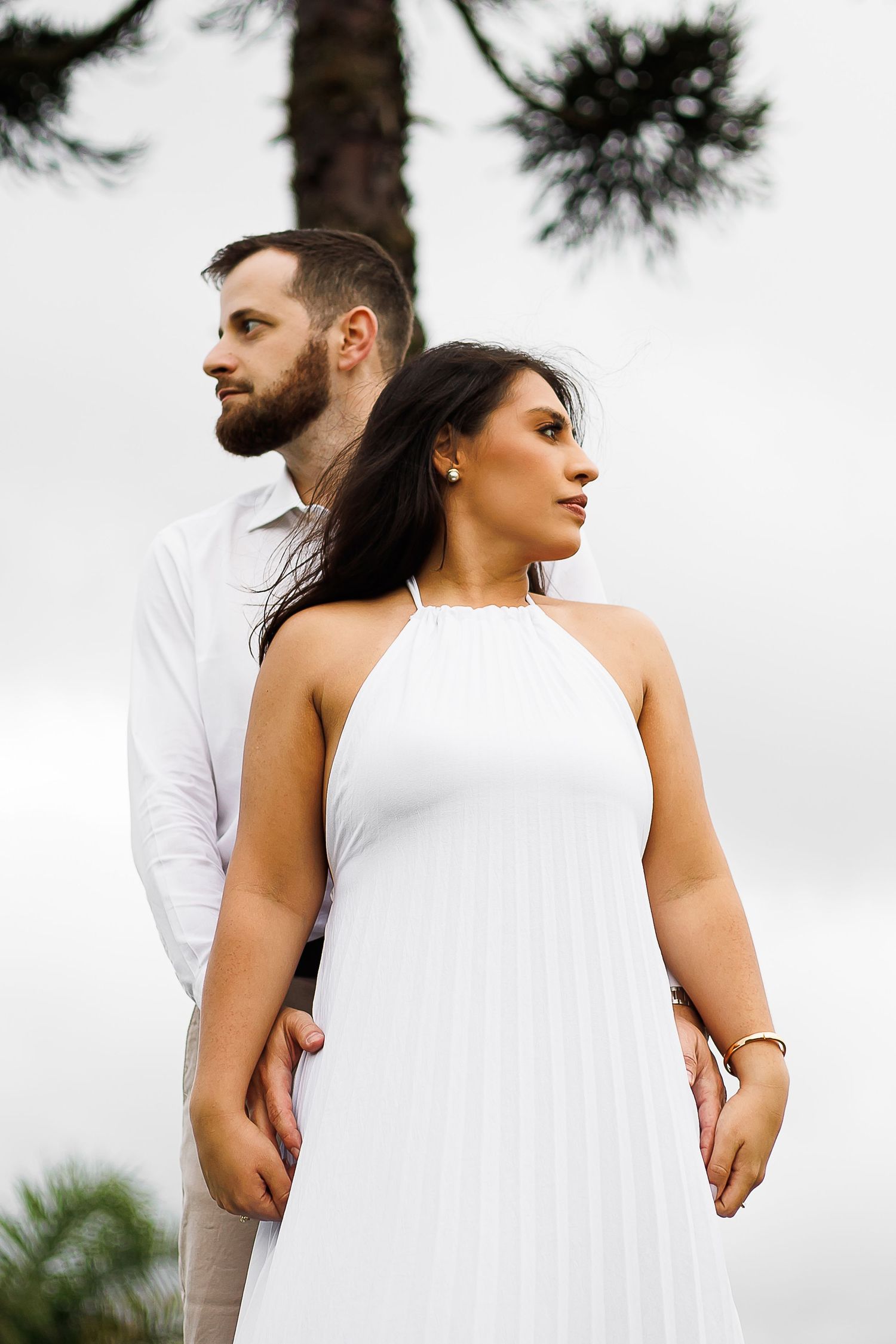 Couple in white attire standing back to back outdoors, woman in halter dress, bearded man in shirt, pine tree background.