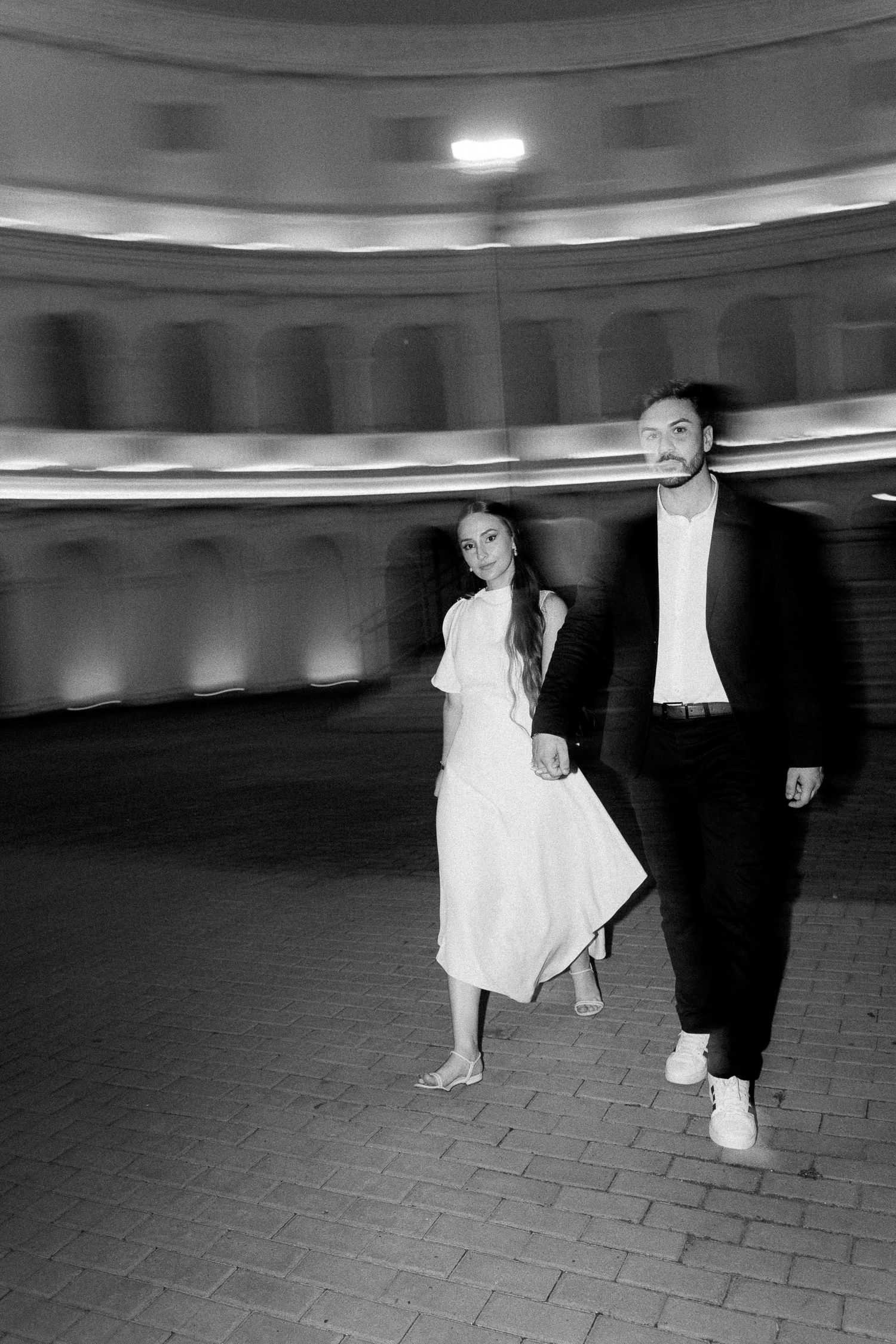 Black and white photo of a couple walking hand in hand at night near an illuminated classical building.