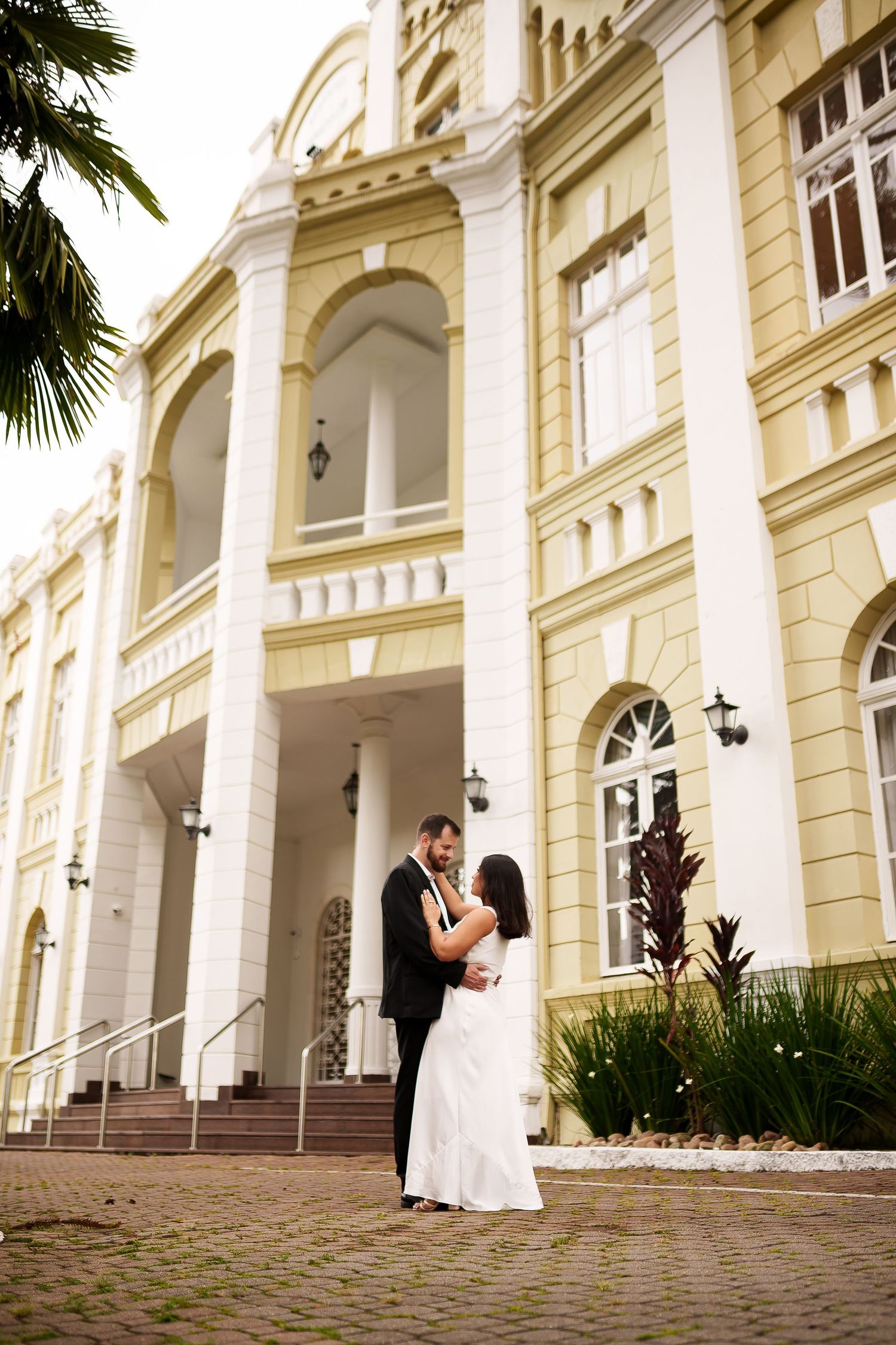 Bride and groom embrace in front of elegant yellow colonial building with white arches and steps.