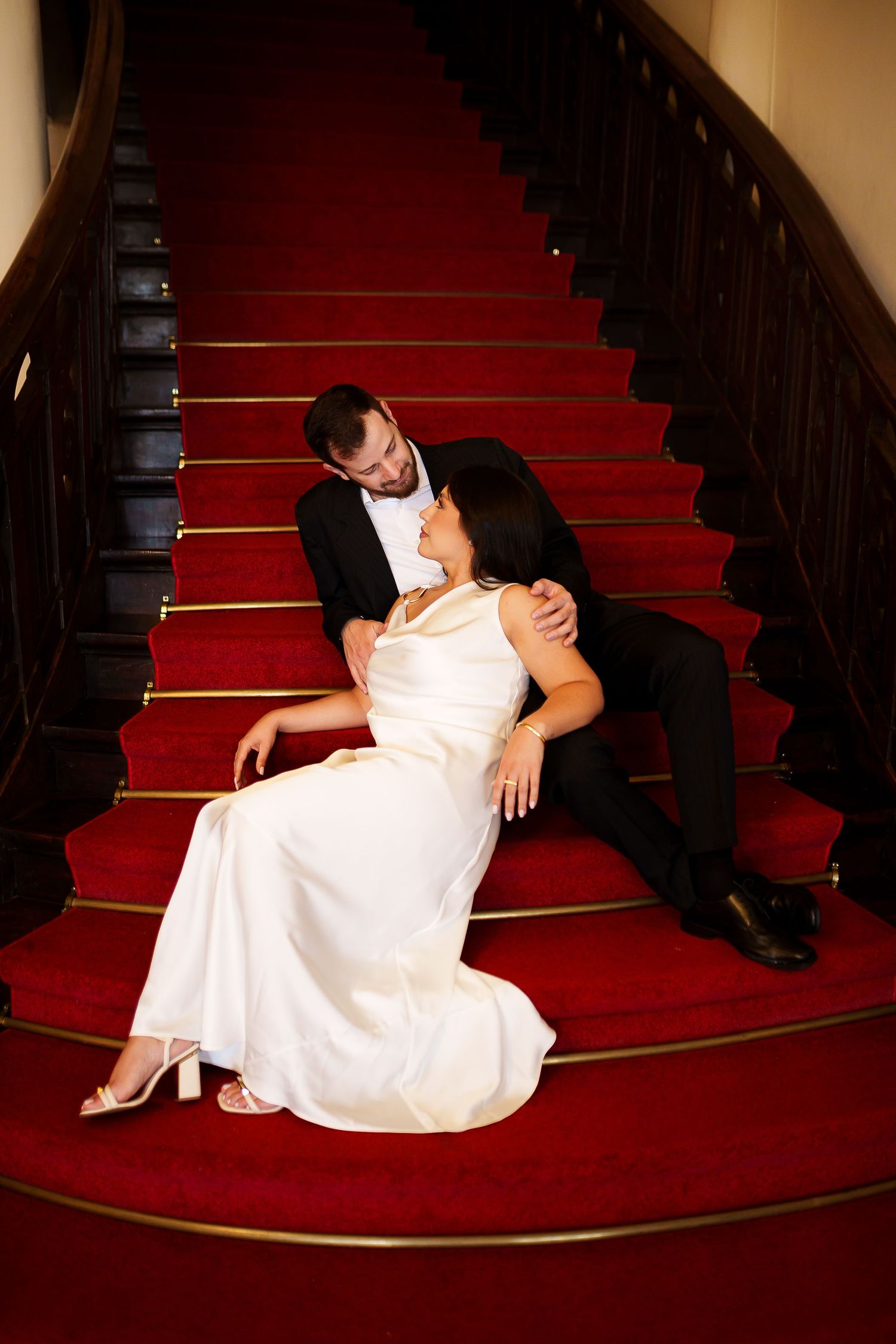 Bride in white gown and groom in black tuxedo sitting together on red carpeted staircase in romantic pose.
