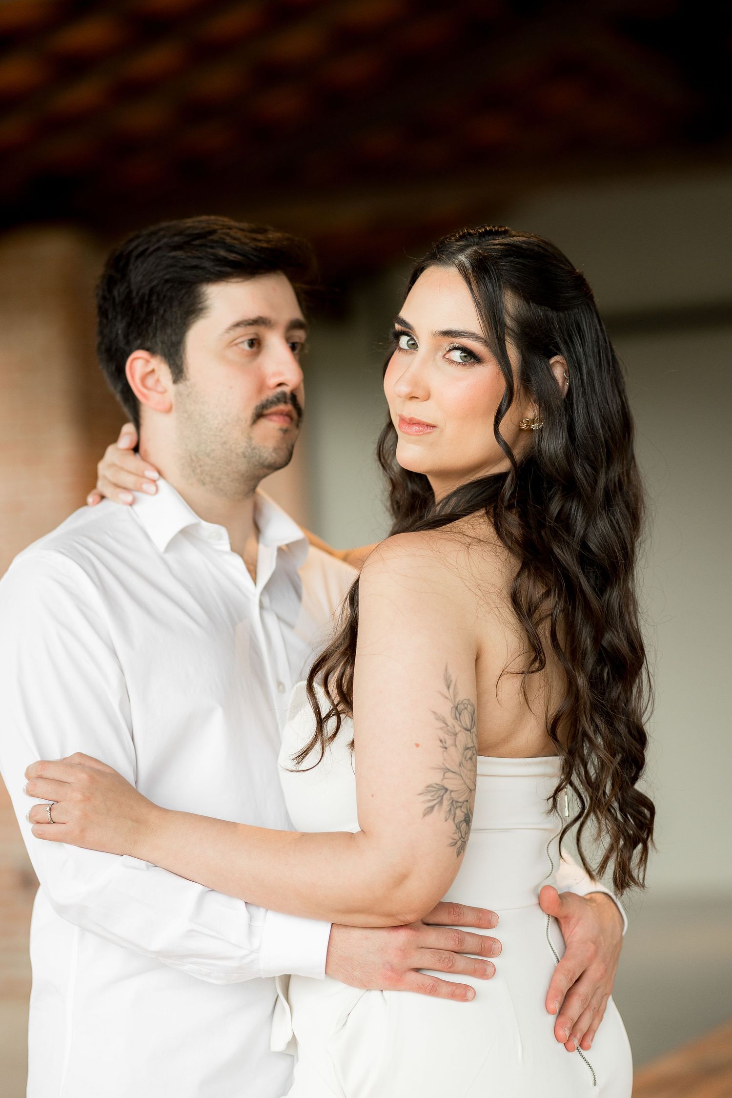 Couple in white attire embracing outdoors, woman with long wavy hair and floral arm tattoo gazing at camera.