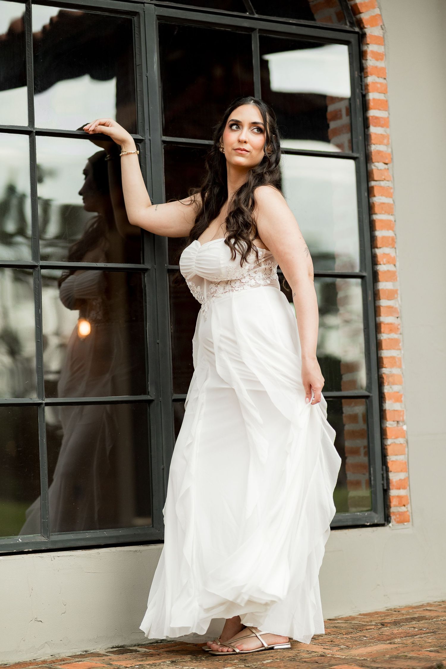 Bride in flowing white strapless gown poses gracefully by large black-framed windows with brick wall backdrop.