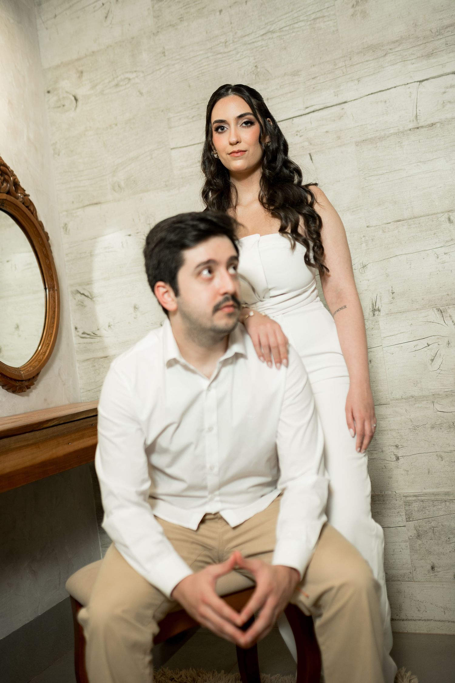 Elegant couple in white attire posing in a studio, woman standing behind seated man near a vintage mirror.
