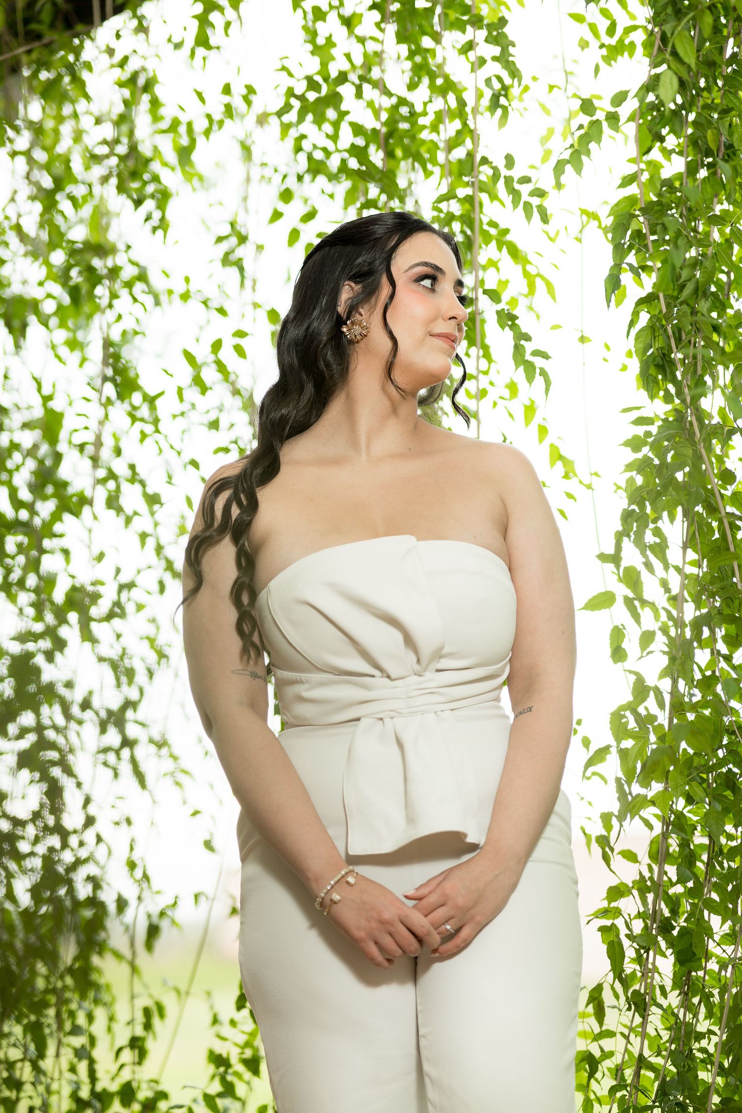Woman in white strapless jumpsuit posing among lush green hanging leaves, wearing gold jewelry.