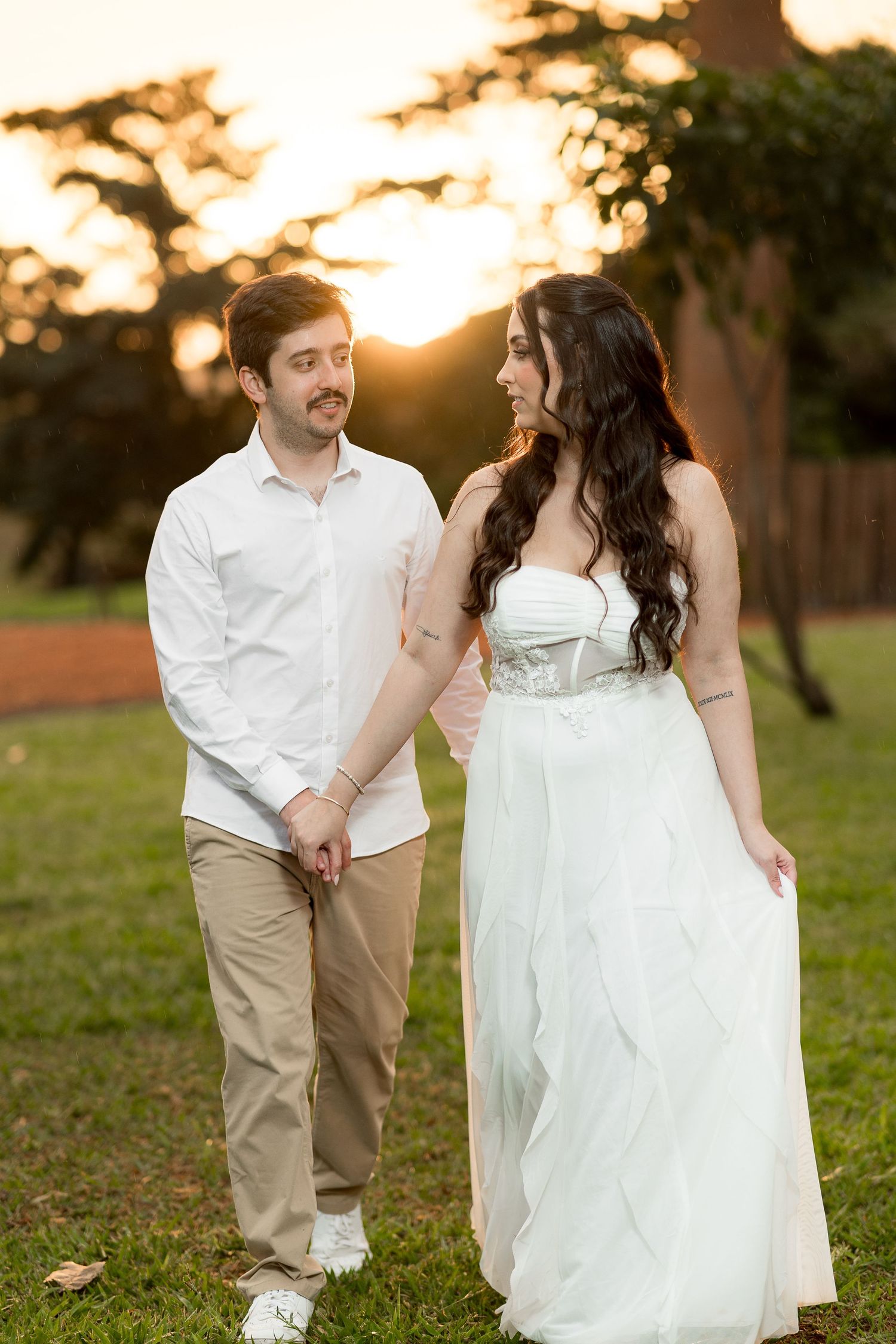 Couple holding hands walking in park at golden sunset, woman in white bridal gown, man in white shirt and khaki pants.