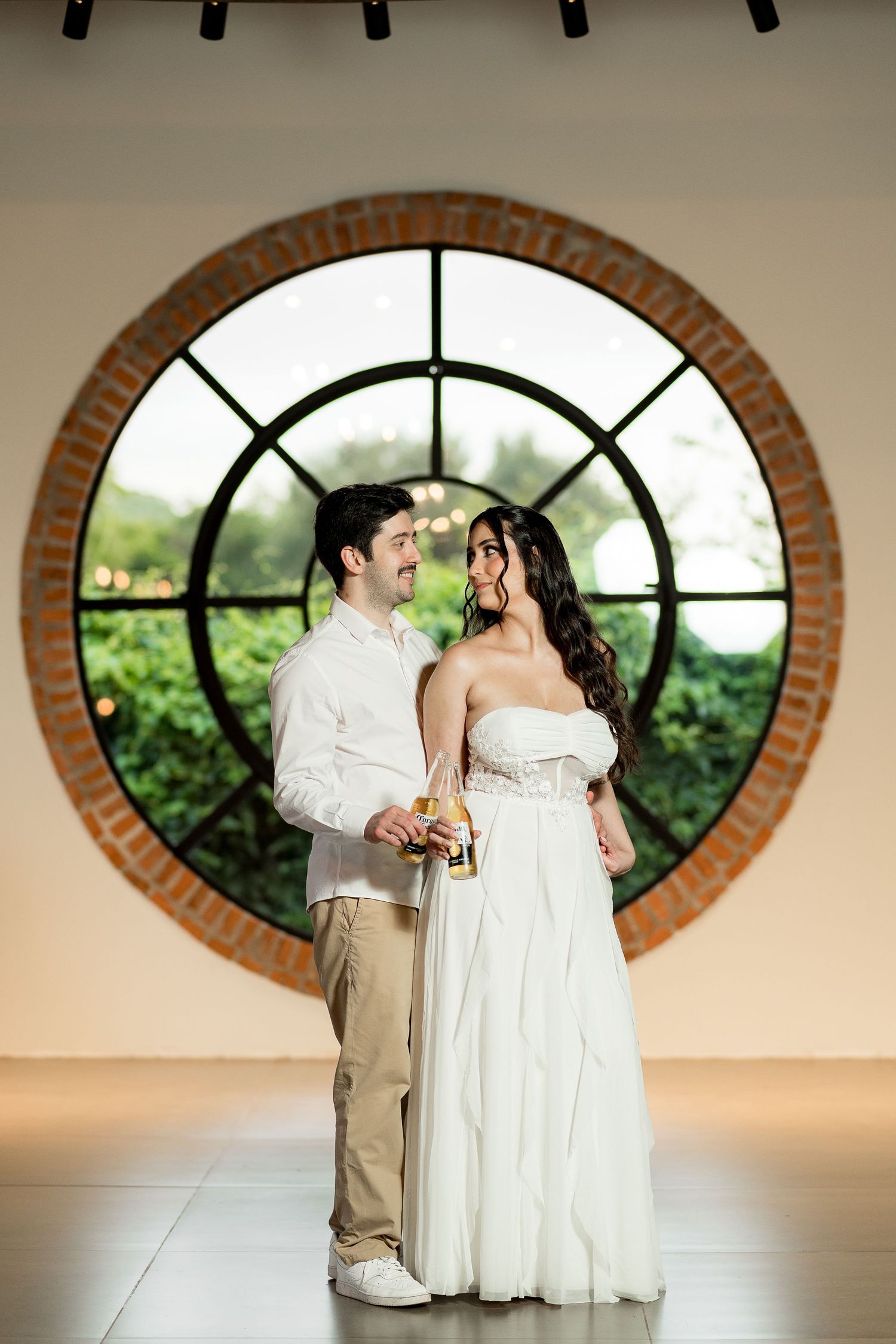 Couple in wedding attire embracing before a round brick-framed window with lush greenery backdrop.